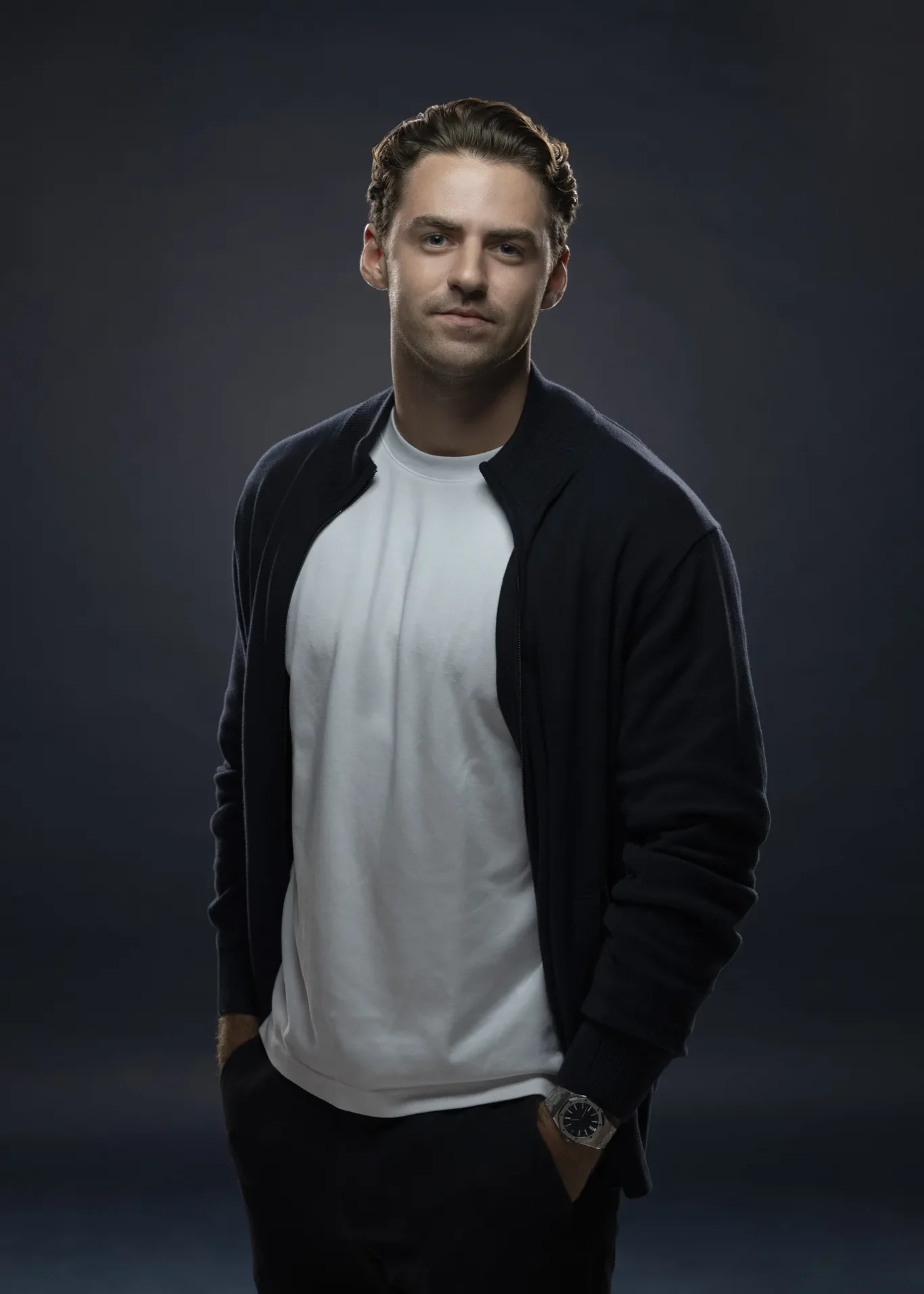 Portrait of a man in a white t-shirt and dark cardigan, with hands in pockets, wearing a silver watch, against a dark gradient backdrop.