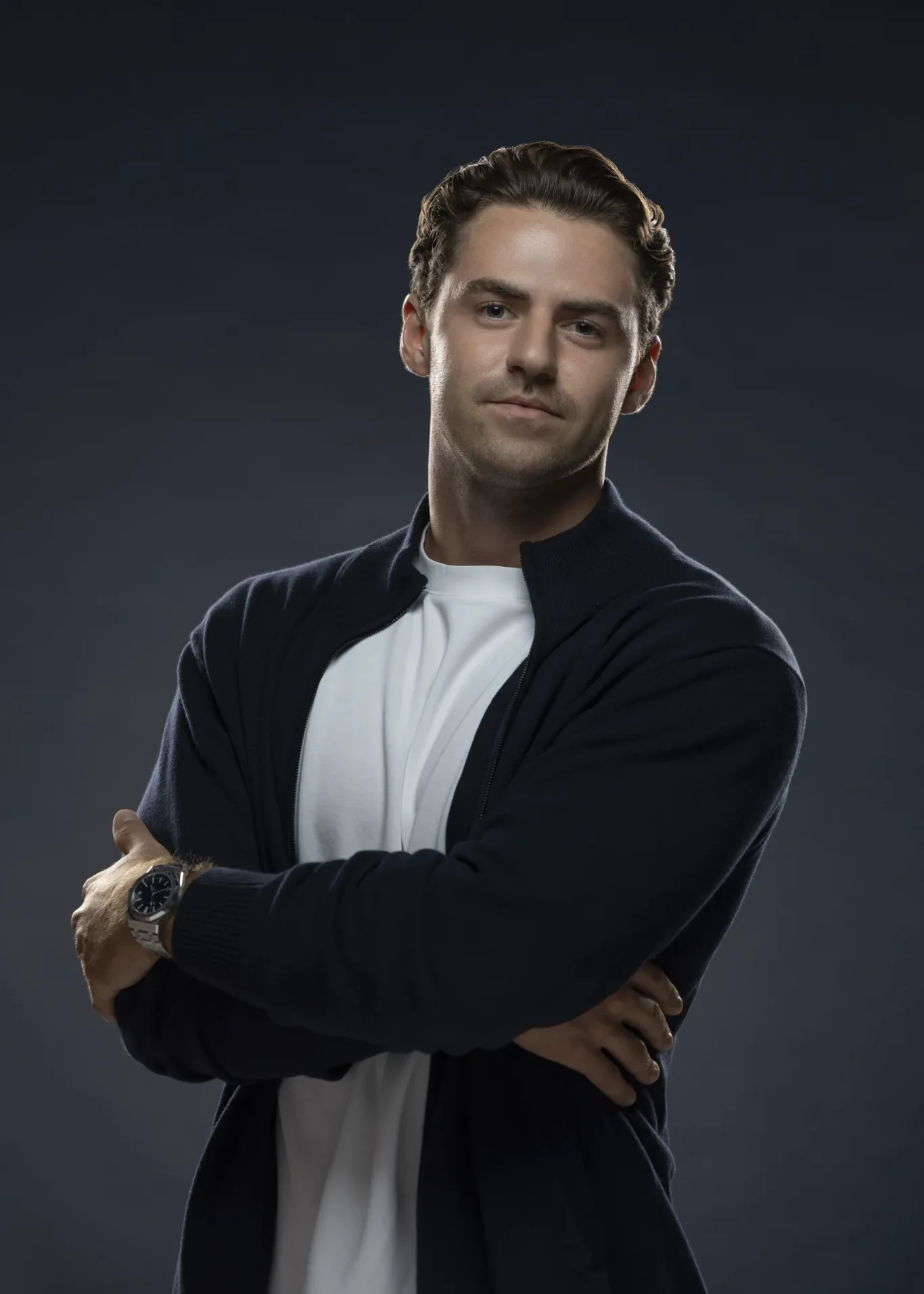 Portrait of a man with arms crossed, wearing a white shirt and dark cardigan against a dark studio background.