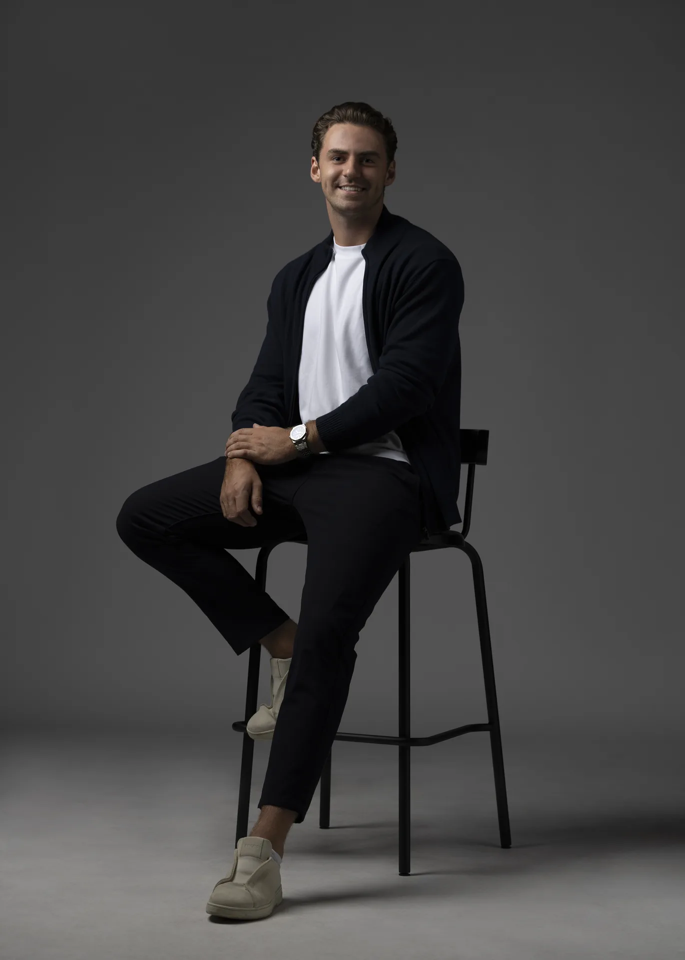 Smiling man seated on a tall stool in a studio, wearing a white shirt, dark cardigan, light sneakers, against a gray backdrop.