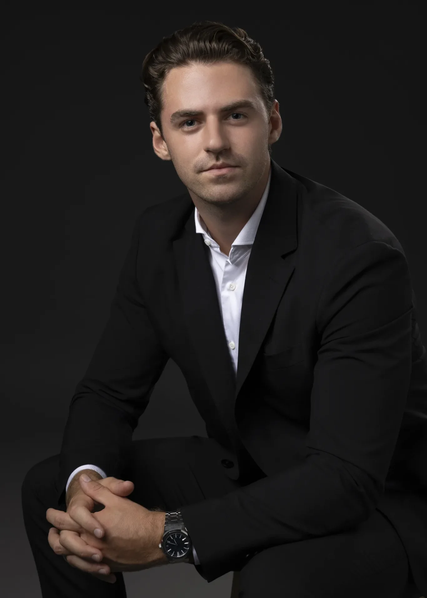 Portrait of a man in a black suit and white shirt, seated with hands clasped, looking at the camera against a dark background.