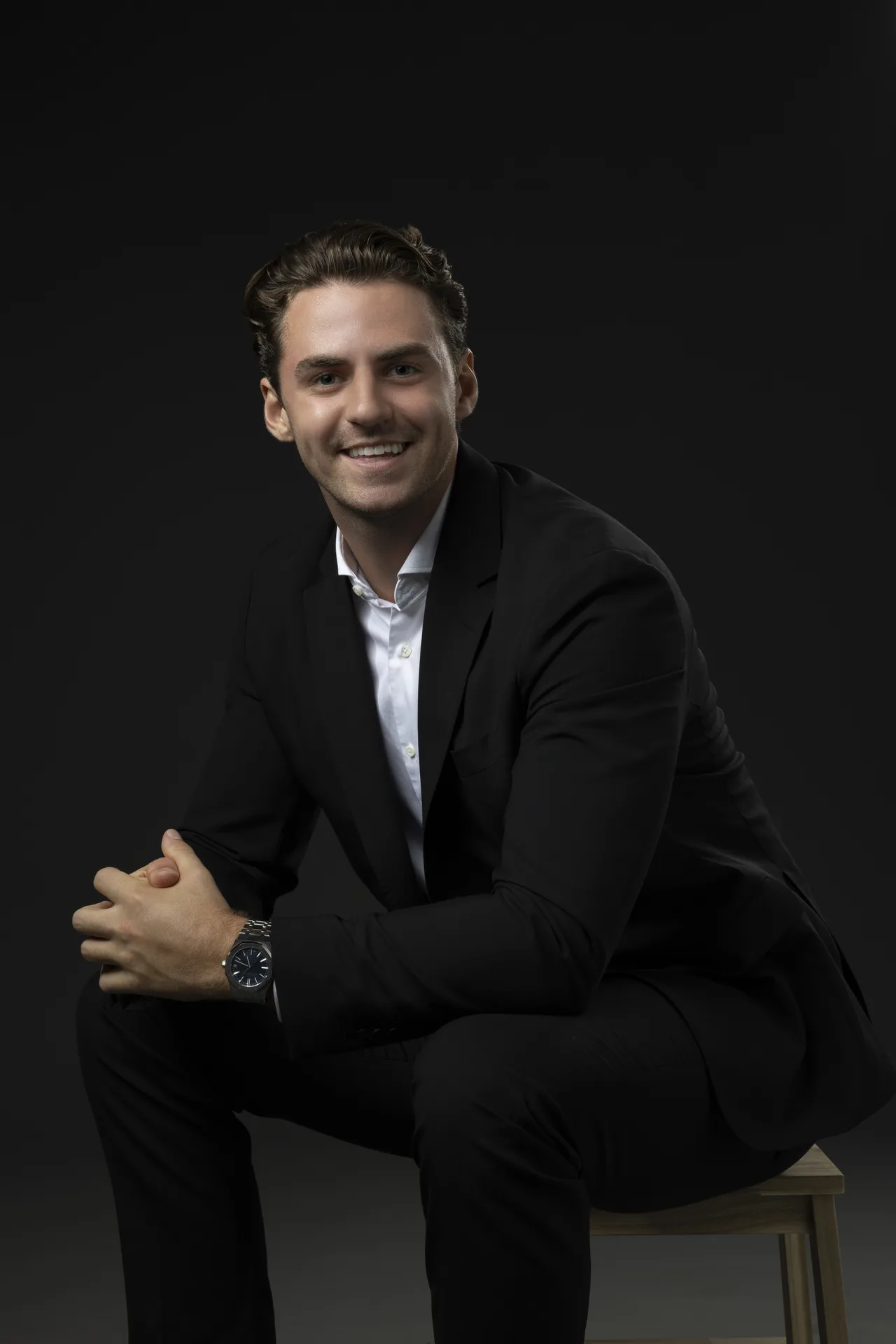 Smiling man in a black suit sits on a wooden bench against a dark background, hands clasped in front.