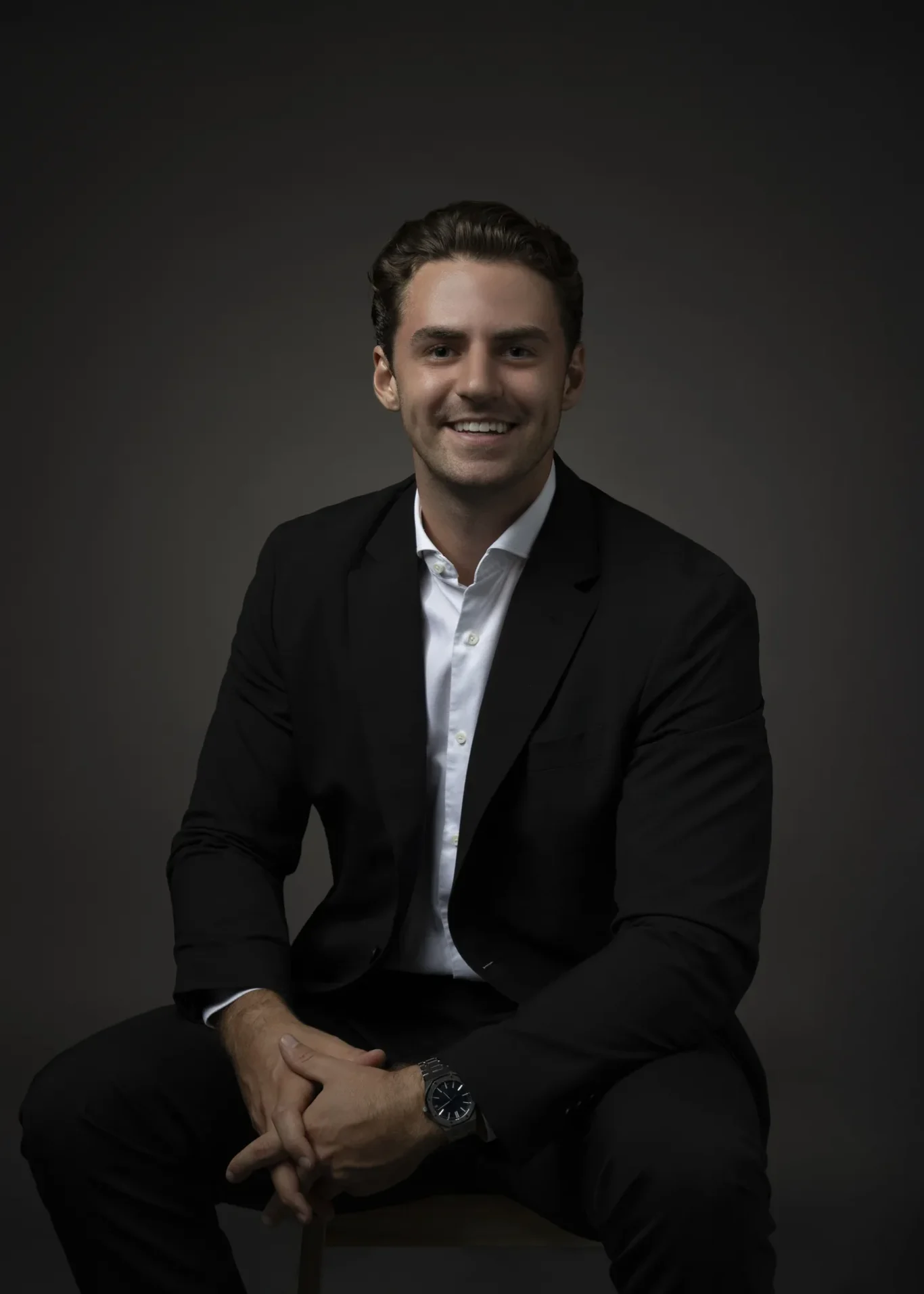 Smiling man in a black suit and white shirt seated against a dark gradient background.