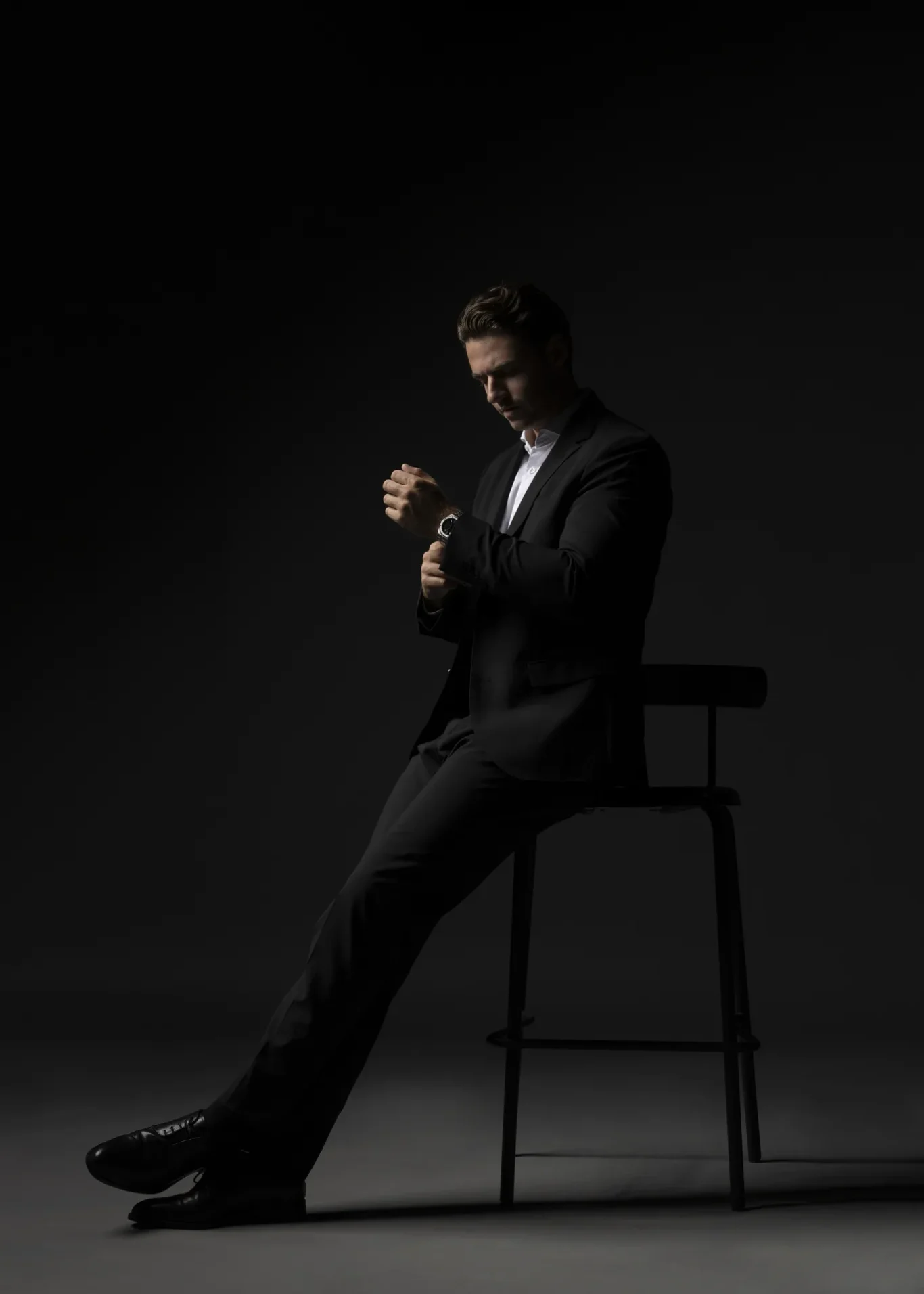 Man in a dark suit sits on a tall stool, adjusting his wristwatch in a dim studio.
