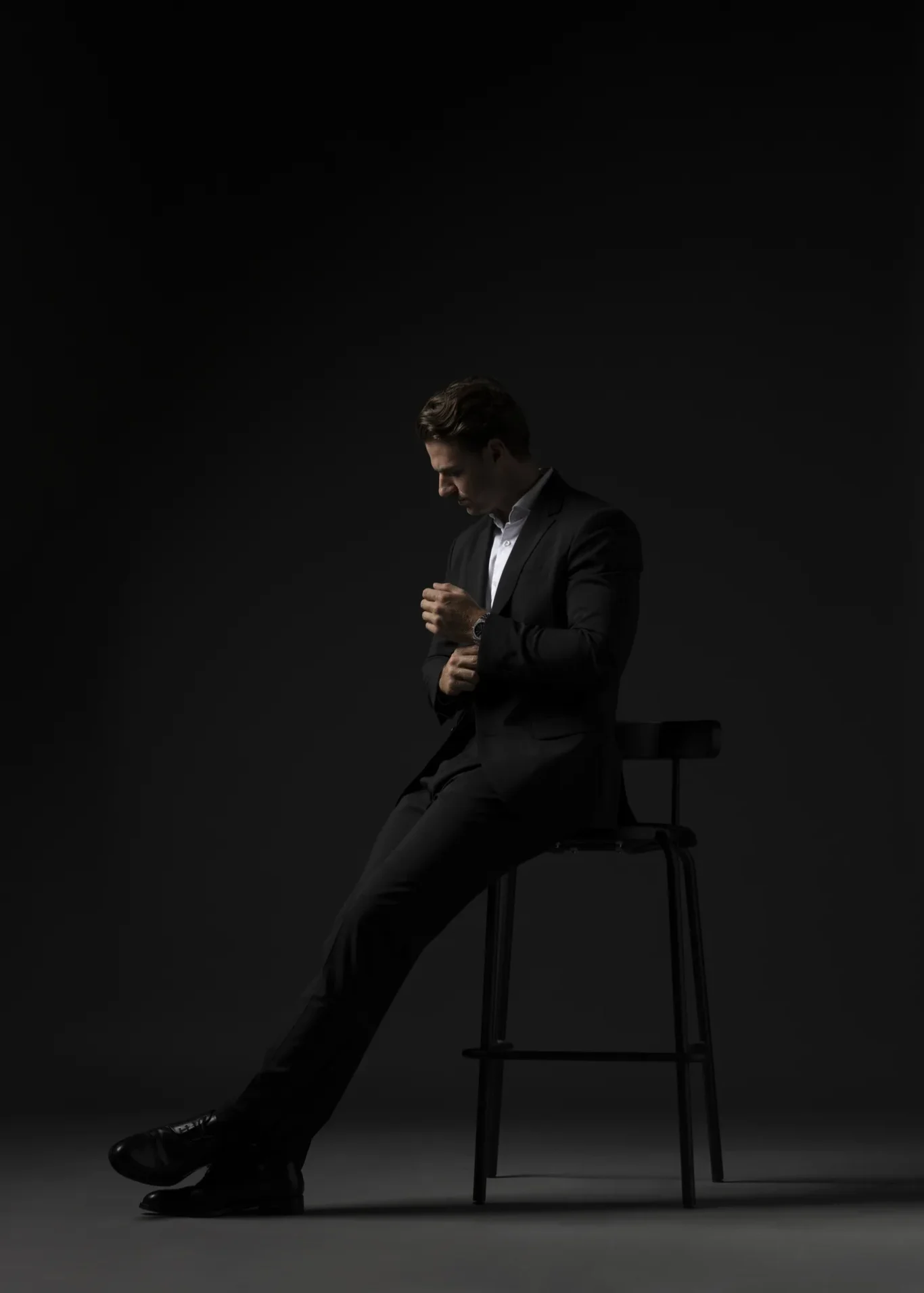 Man in a dark suit sits on a tall stool, head bowed, adjusting his cuff against a black studio background.