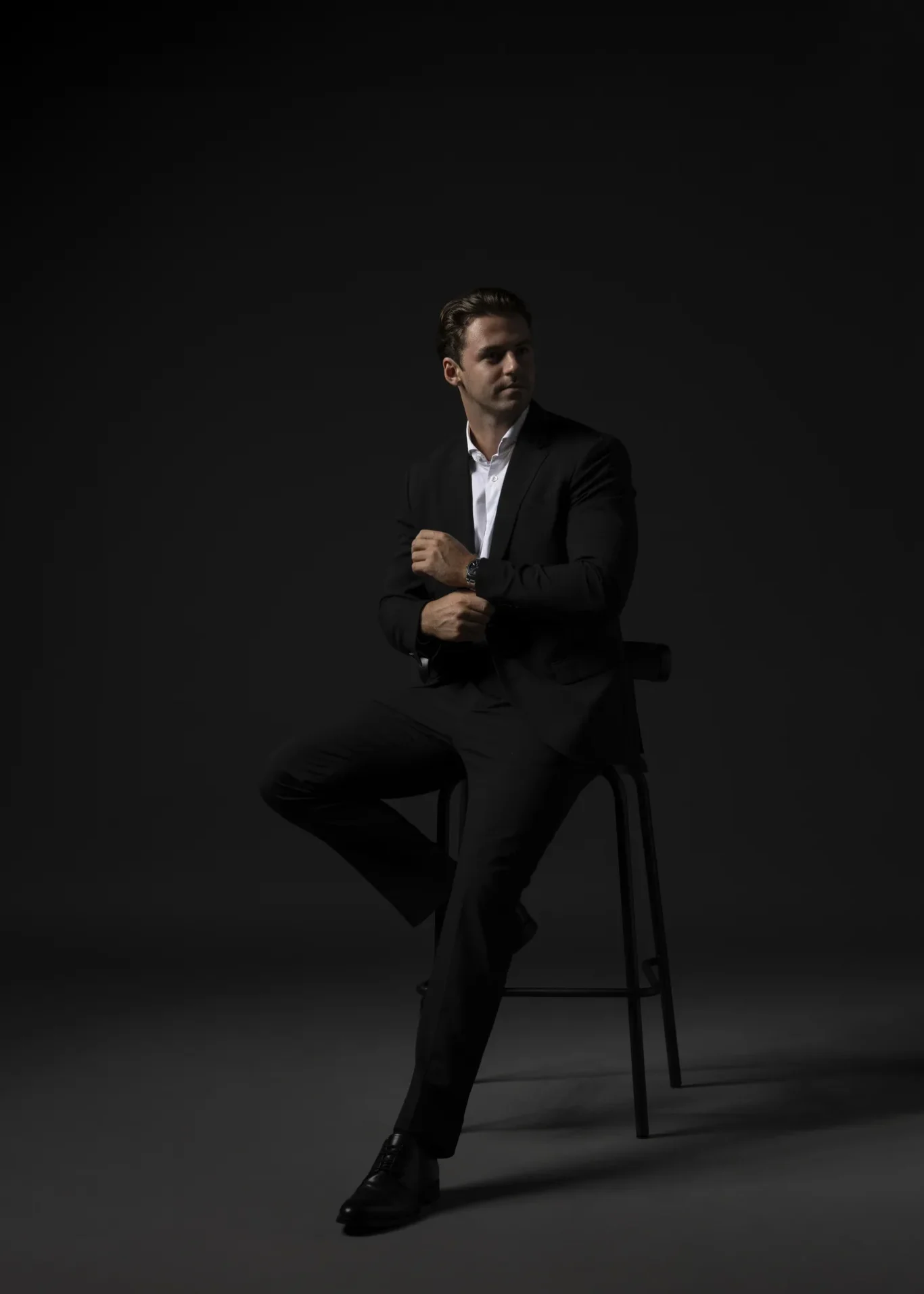 Man in a black suit seated on a tall stool in a dark studio, looking to the right.