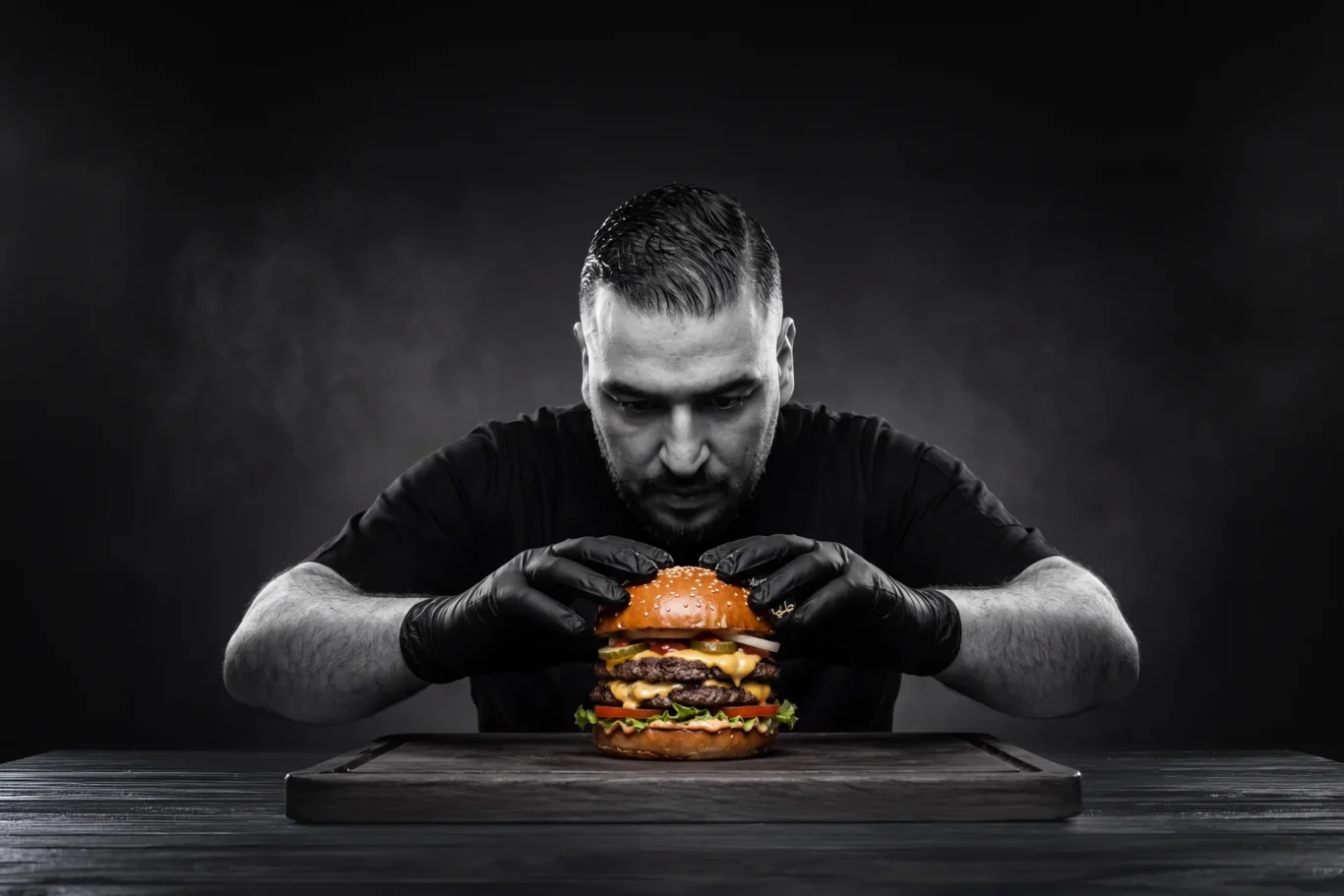 Chef Abdul Rahman Mustapha in black gloves pressing a double cheeseburger on a wooden board, a black and white image with the burger in colour, created by MRPS with AI.
