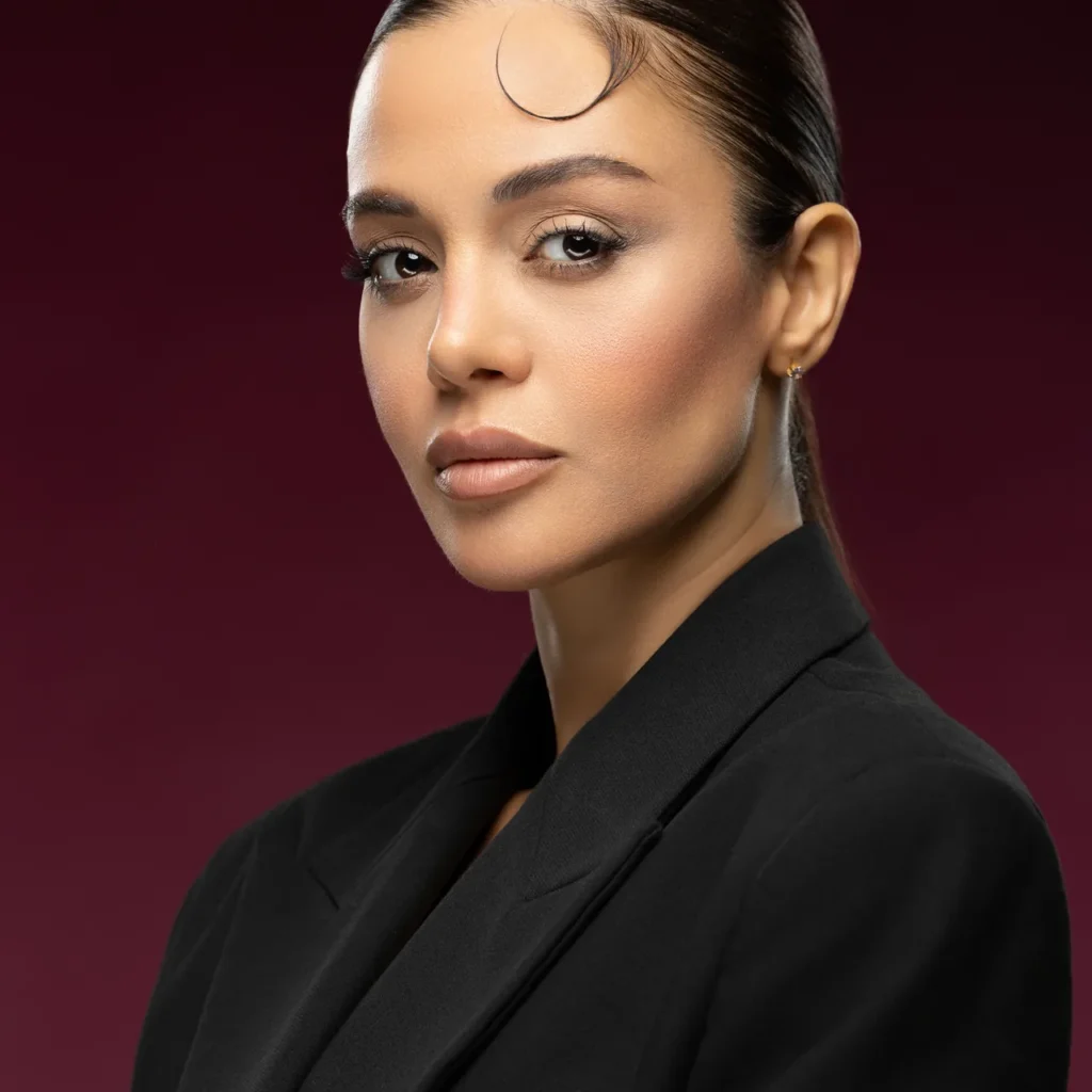 Close-up professional headshot of a businesswoman in a black blazer, looking over her shoulder against a deep burgundy studio backdrop.
