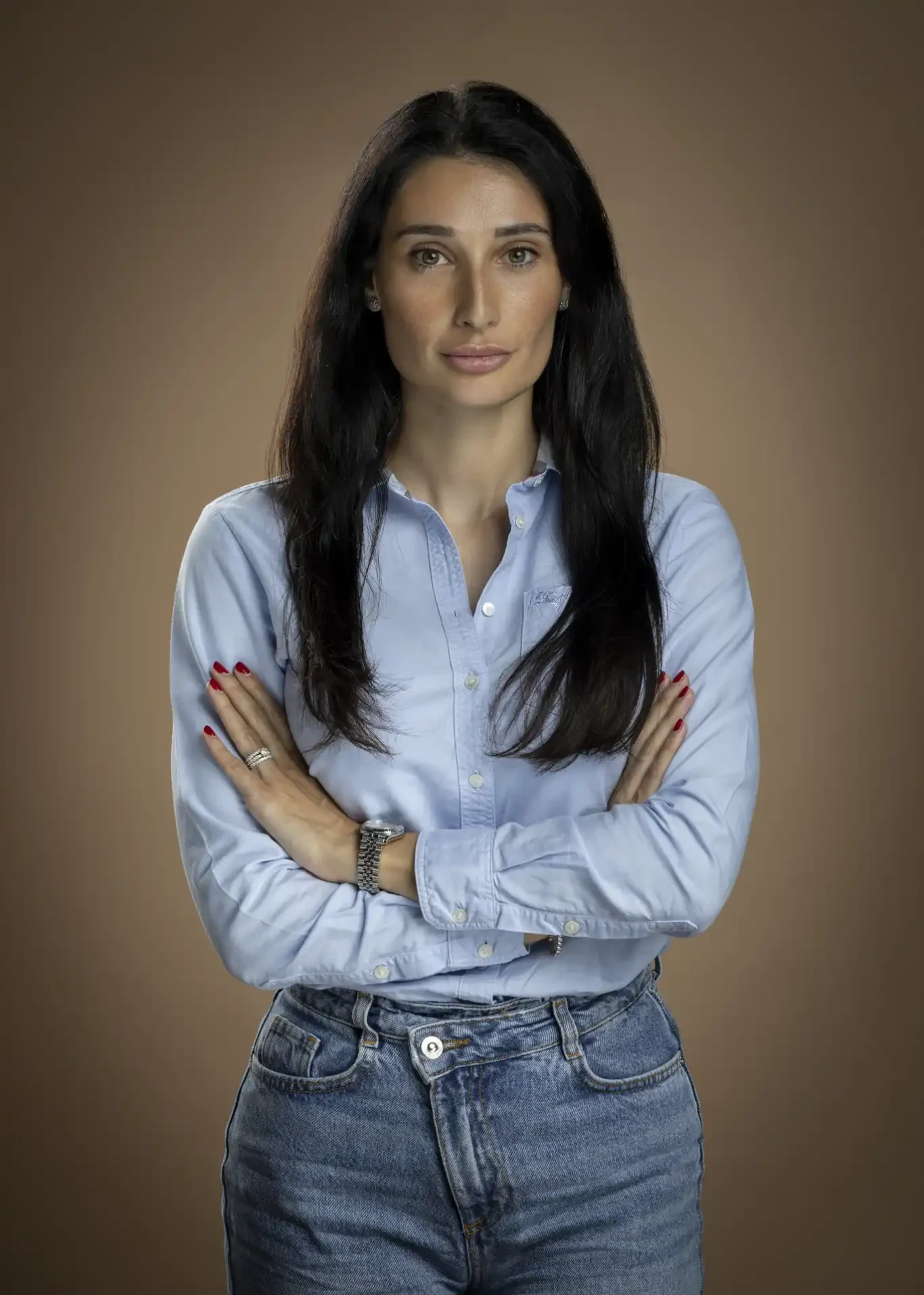 Corporate headshot of a woman in a light blue shirt and jeans with arms crossed, against a warm brown studio backdrop.