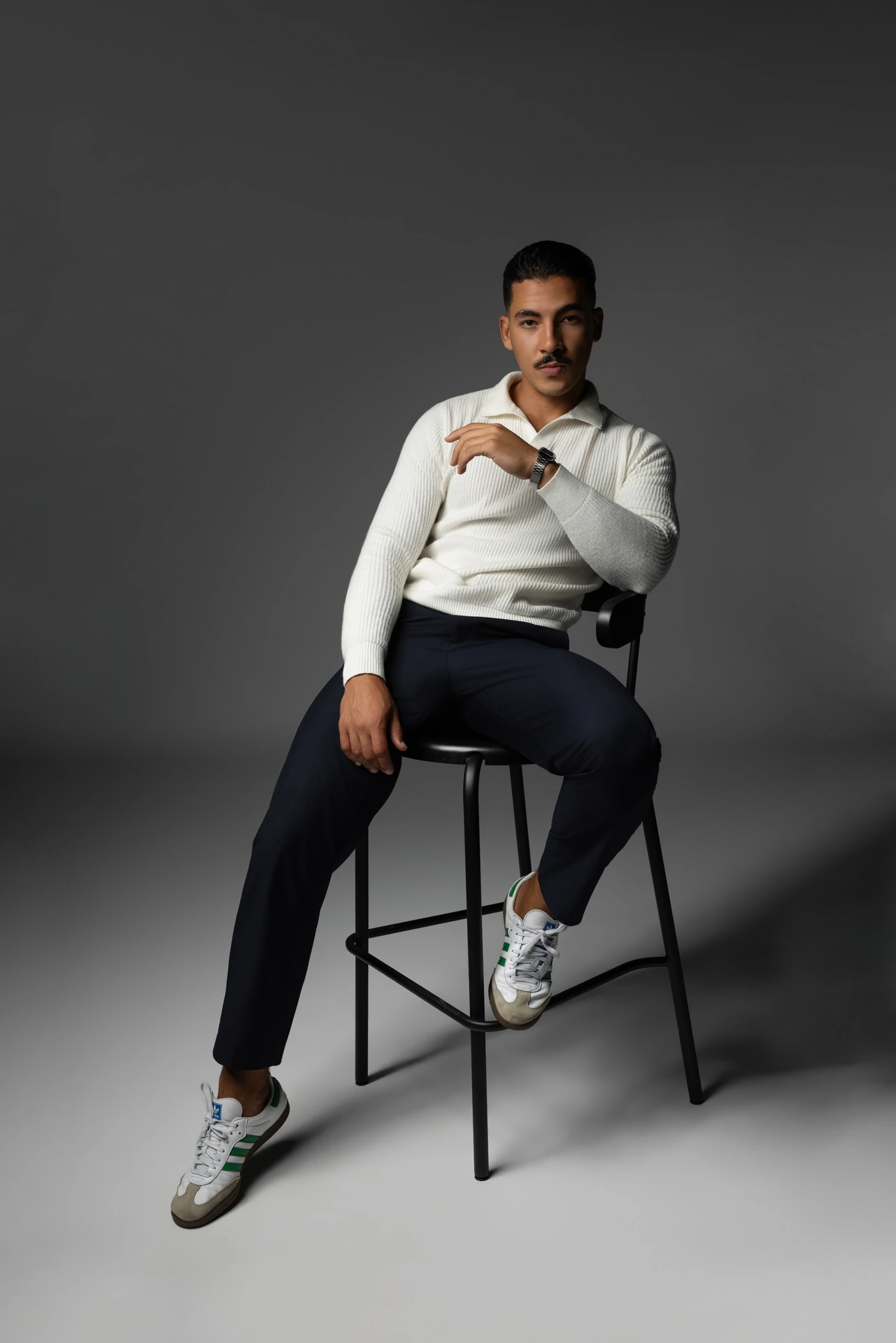 Abdelhak Benatia seated on a stool in a cream knit polo and navy trousers, full length on a grey backdrop. Male modeling portfolio photoshoot by MRPS in Dubai, photographed by Nastaran.
