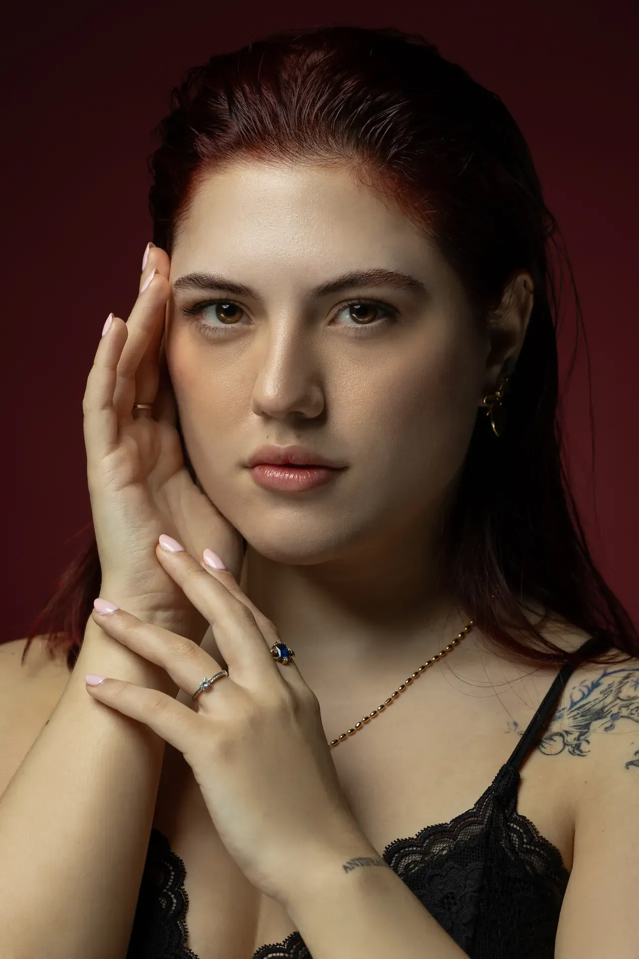 Close beauty portrait with both hands framing the face on a deep burgundy backdrop. Beauty portrait of model Julian, photographed by Nastaran at the MRPS studio in Dubai.