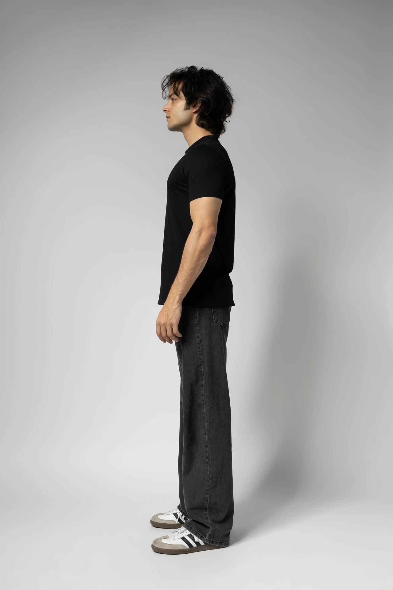 Full-length side view of male model Arsh in a black t-shirt, dark jeans and sneakers on a light grey studio backdrop.
