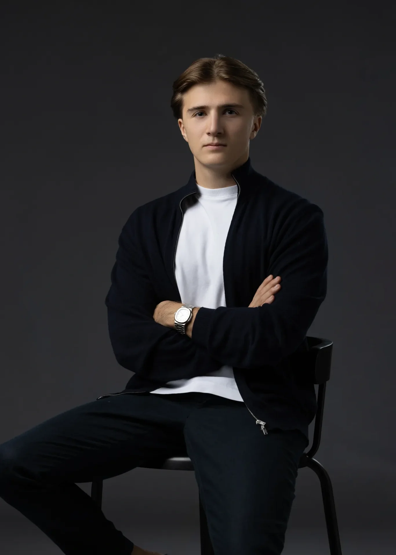 Young entrepreneur seated on a black chair with arms crossed, navy cardigan over a white t-shirt, dark grey backdrop. Editorial business portrait by MRPS in Dubai for a financial magazine feature.