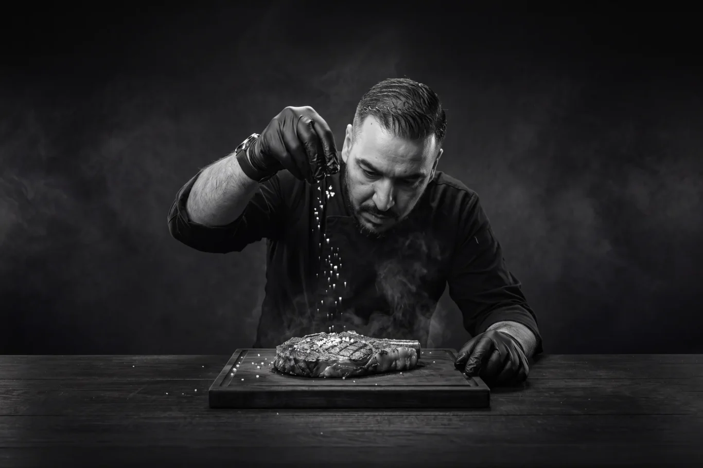 Chef Abdul Rahman Mustapha sprinkling salt over a smoking steak on a wooden board, a black and white AI-assisted image by MRPS.