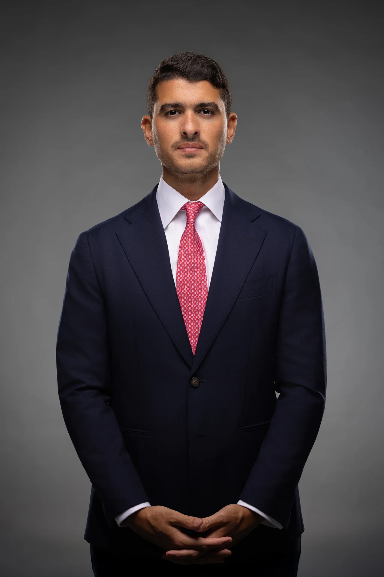 Three-quarter corporate portrait of a business owner in a navy suit and pink tie on a grey studio backdrop.