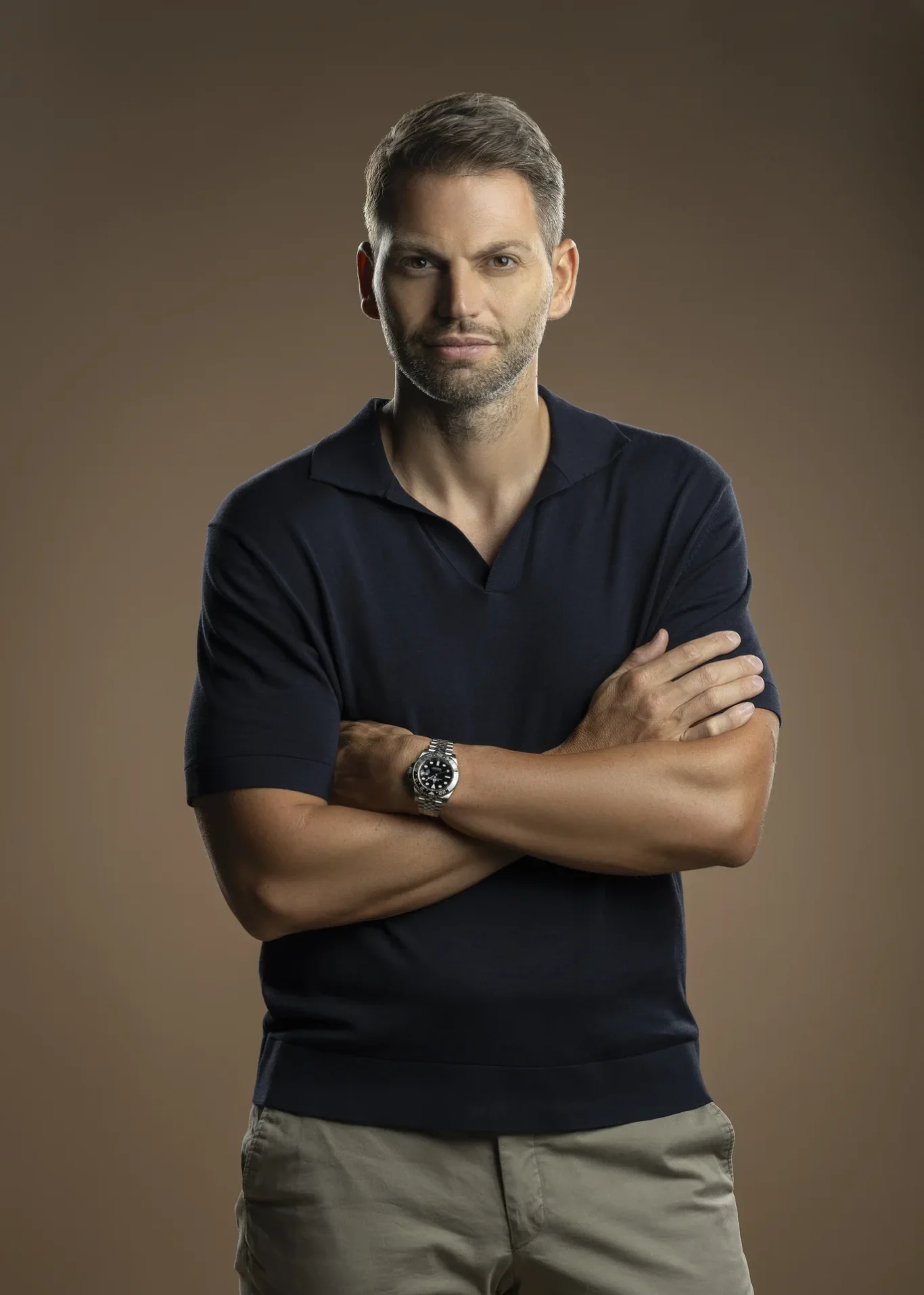 Corporate headshot of a man in a navy polo shirt with arms crossed, against a warm brown studio backdrop.