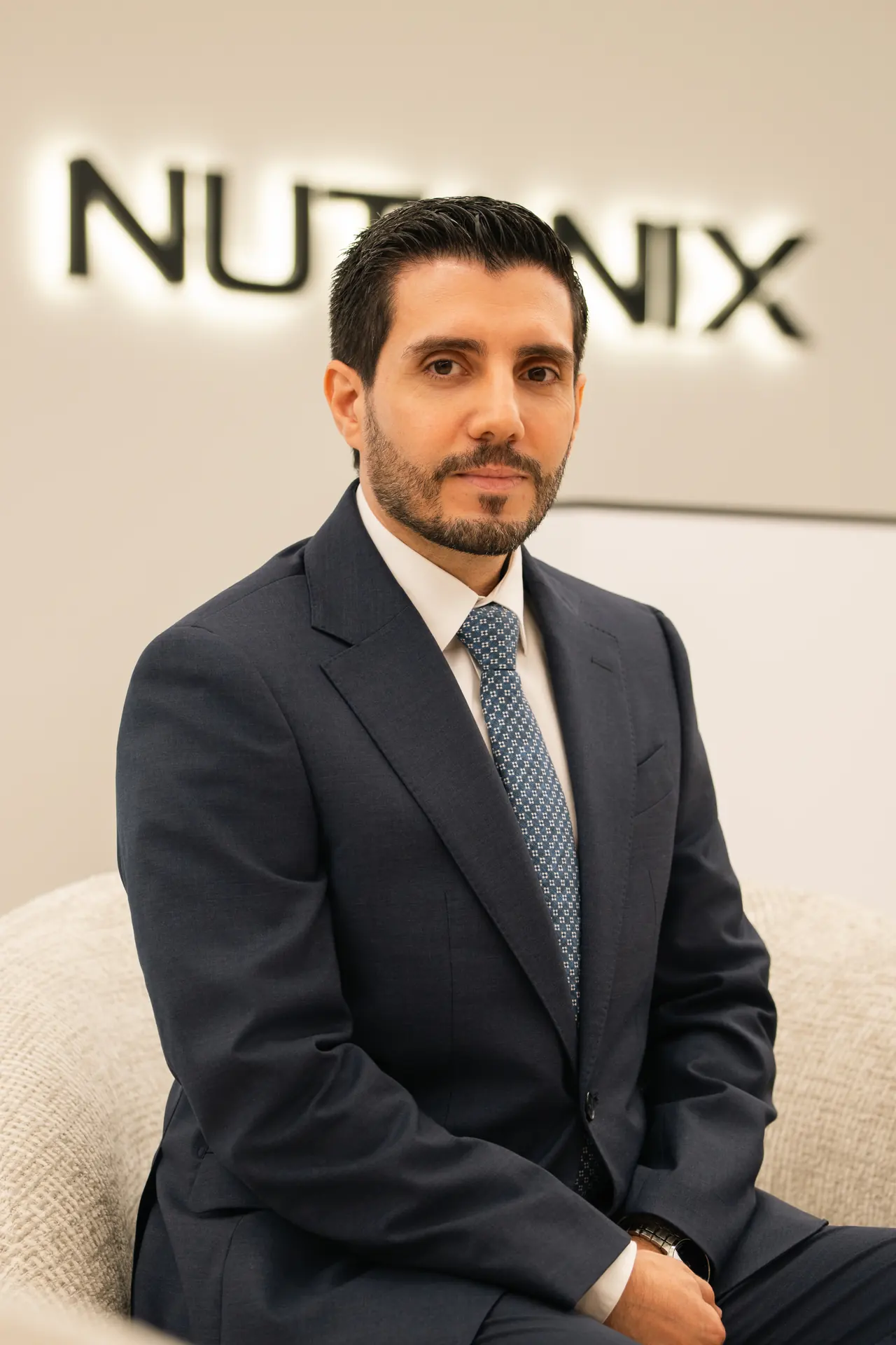 Manager in a navy suit seated in front of the illuminated Nutanix logo. On-site corporate headshot at the Nutanix office in Dubai.