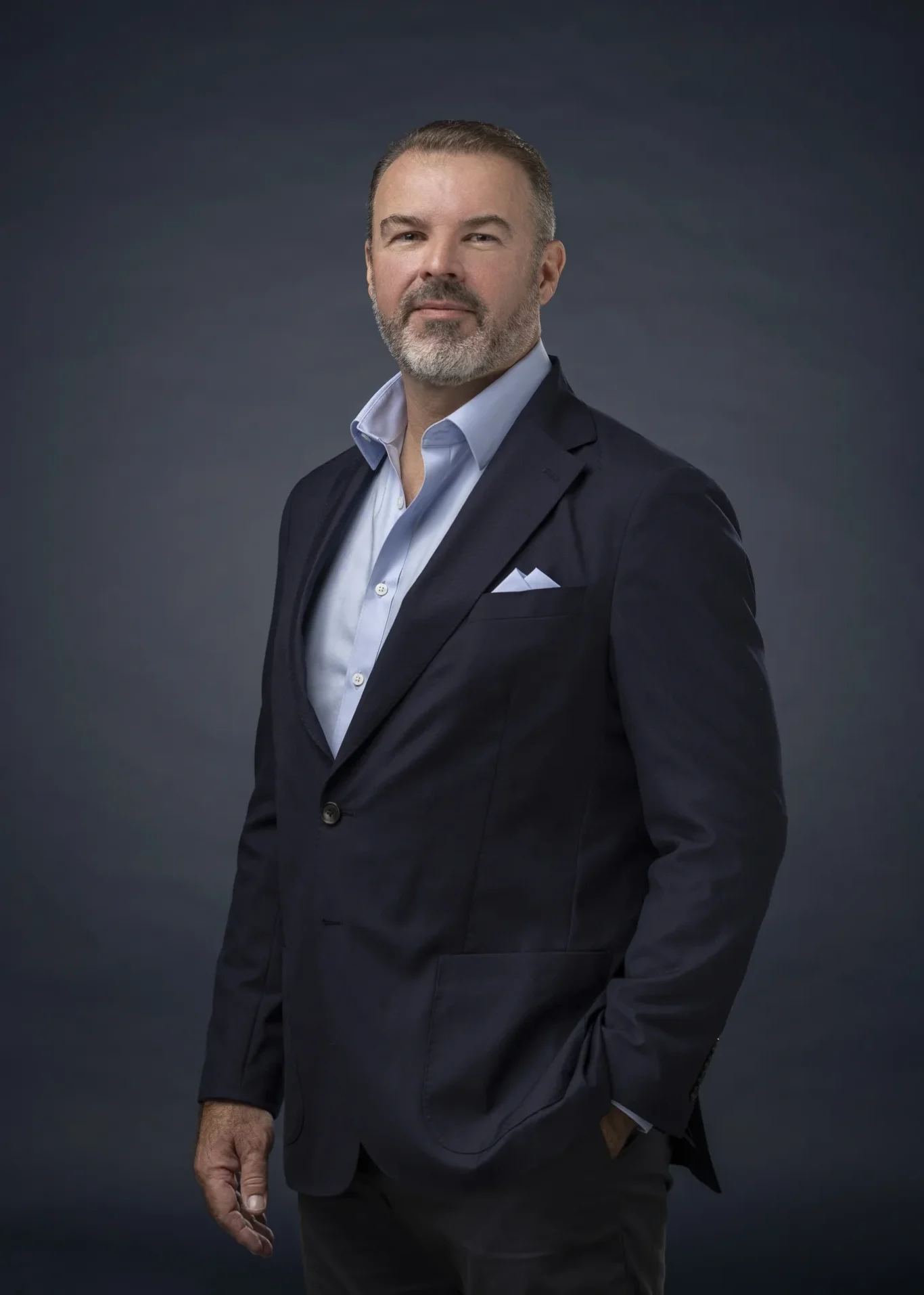 Rik Williams standing in a navy blazer with a pocket square, hand in pocket, dark backdrop. Corporate portrait photography by MRPS in Dubai, photographed by Nastaran.
