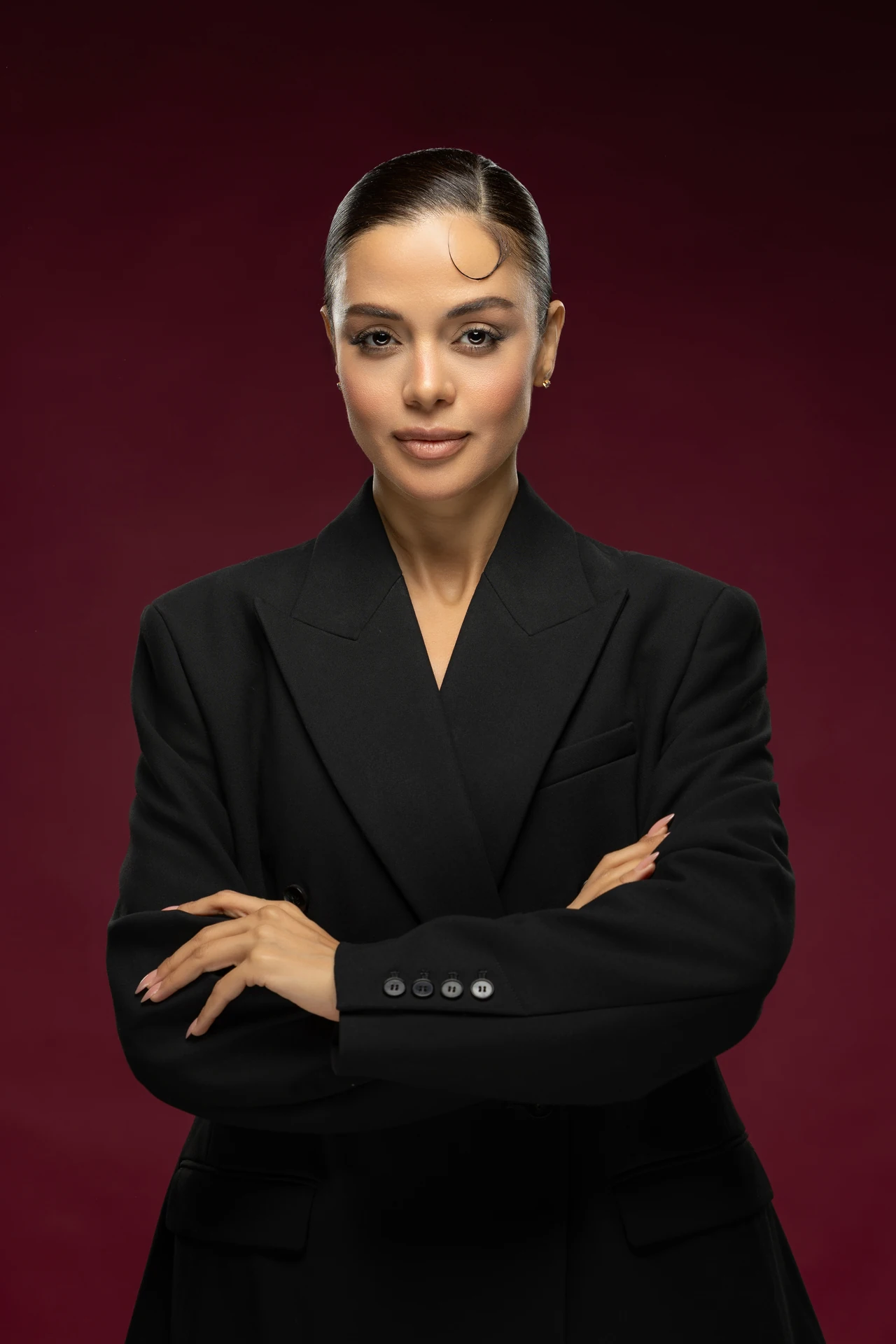Professional headshot of a businesswoman in a black double-breasted blazer with arms crossed, against a deep burgundy studio backdrop.