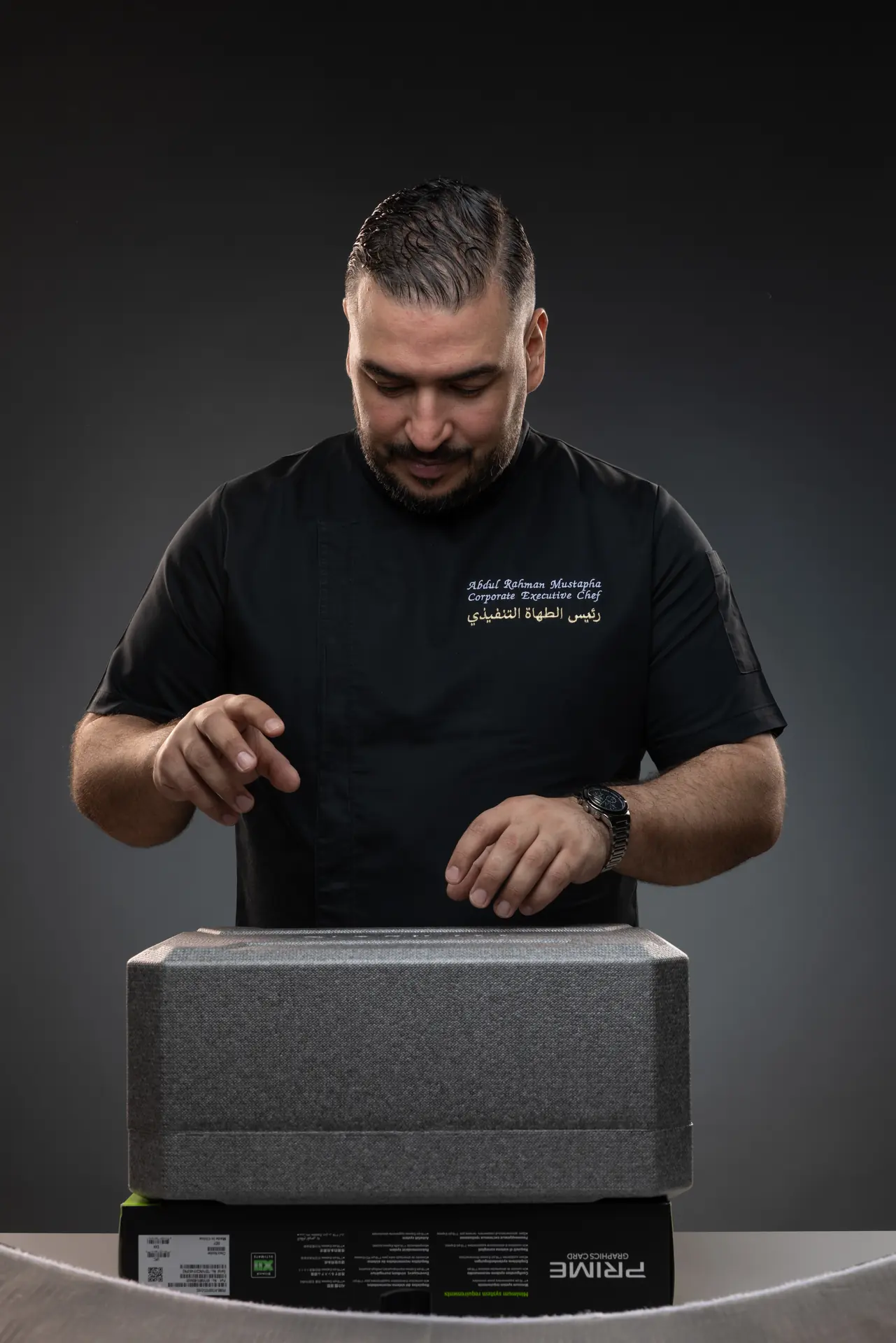 Studio reference photo of the chef posing with both hands over a foam box that stands in for the table.