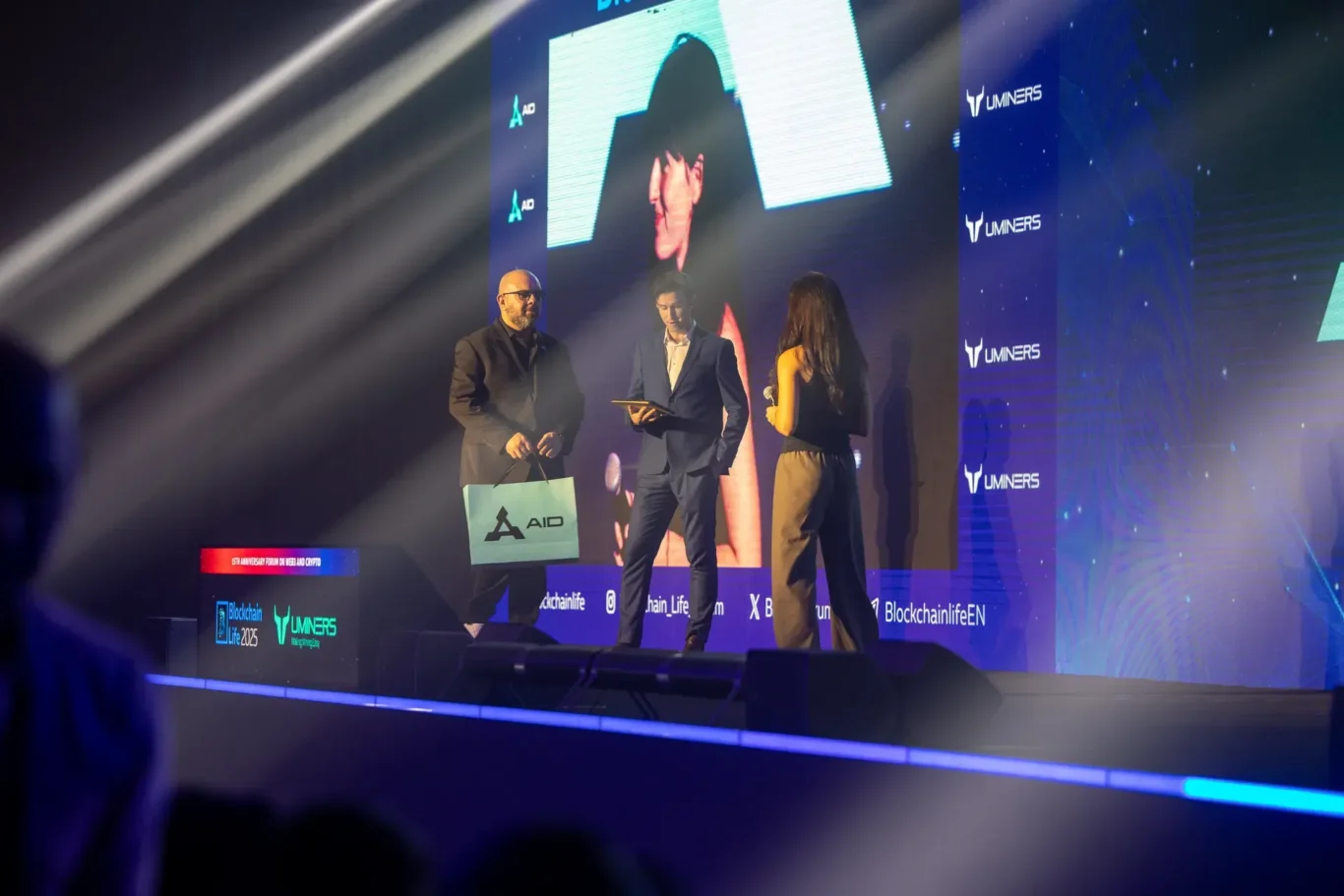 AID representatives on stage in front of the AID logo. Event photography by MRPS at Blockchain Life 2025, Festival Arena, Dubai.
