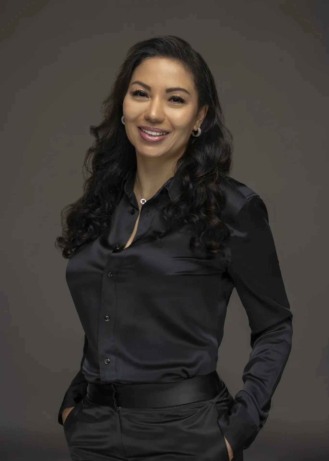 Sandra smiling in a black satin shirt with hands in pockets on a grey backdrop. Business profile and LinkedIn portrait photography by MRPS in Dubai.