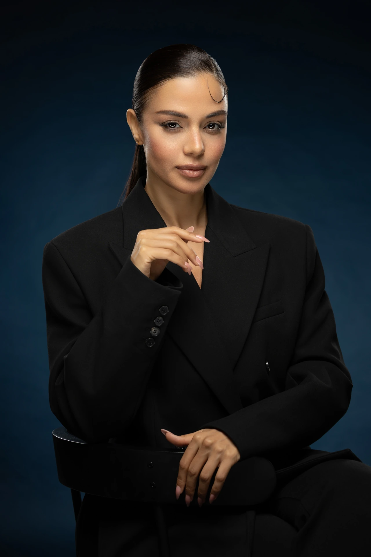 Seated business portrait of a woman in a black suit with one hand raised, against a navy blue studio backdrop.