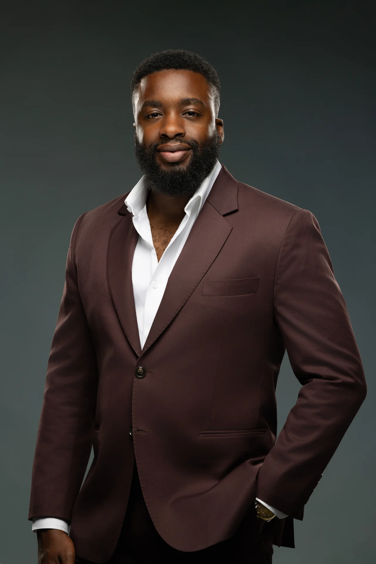 Business owner in a burgundy blazer and white open-collar shirt, hand in pocket, against a dark studio backdrop.