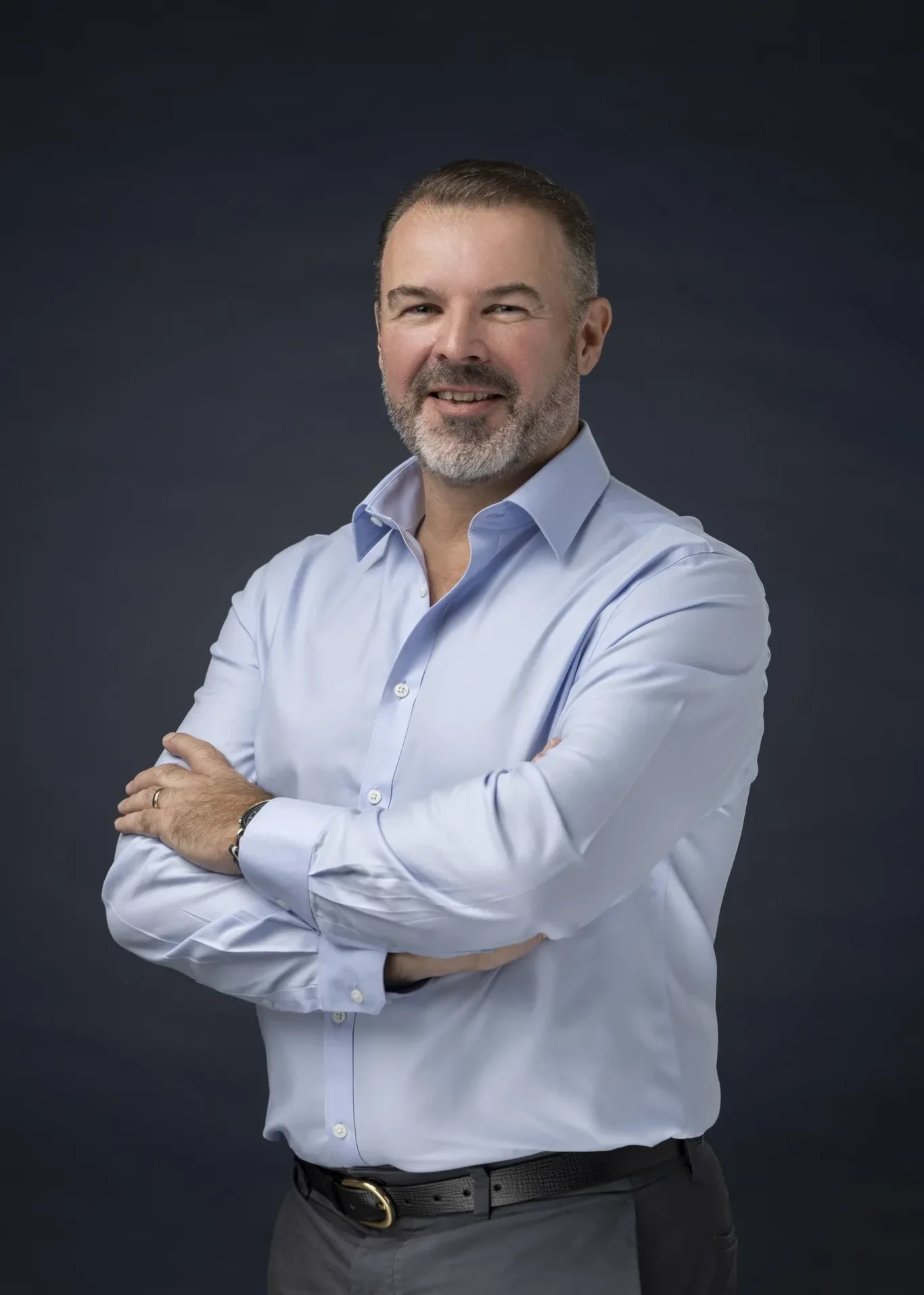 Rik Williams smiling in a light blue shirt, arms crossed, dark navy backdrop. Corporate portrait photography by MRPS in Dubai, photographed by Nastaran.
