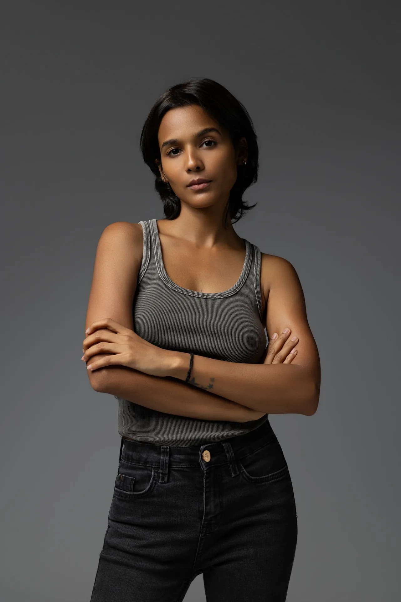 Three-quarter portrait of new face model Mia with arms crossed, grey tank top and black jeans, studio lighting.