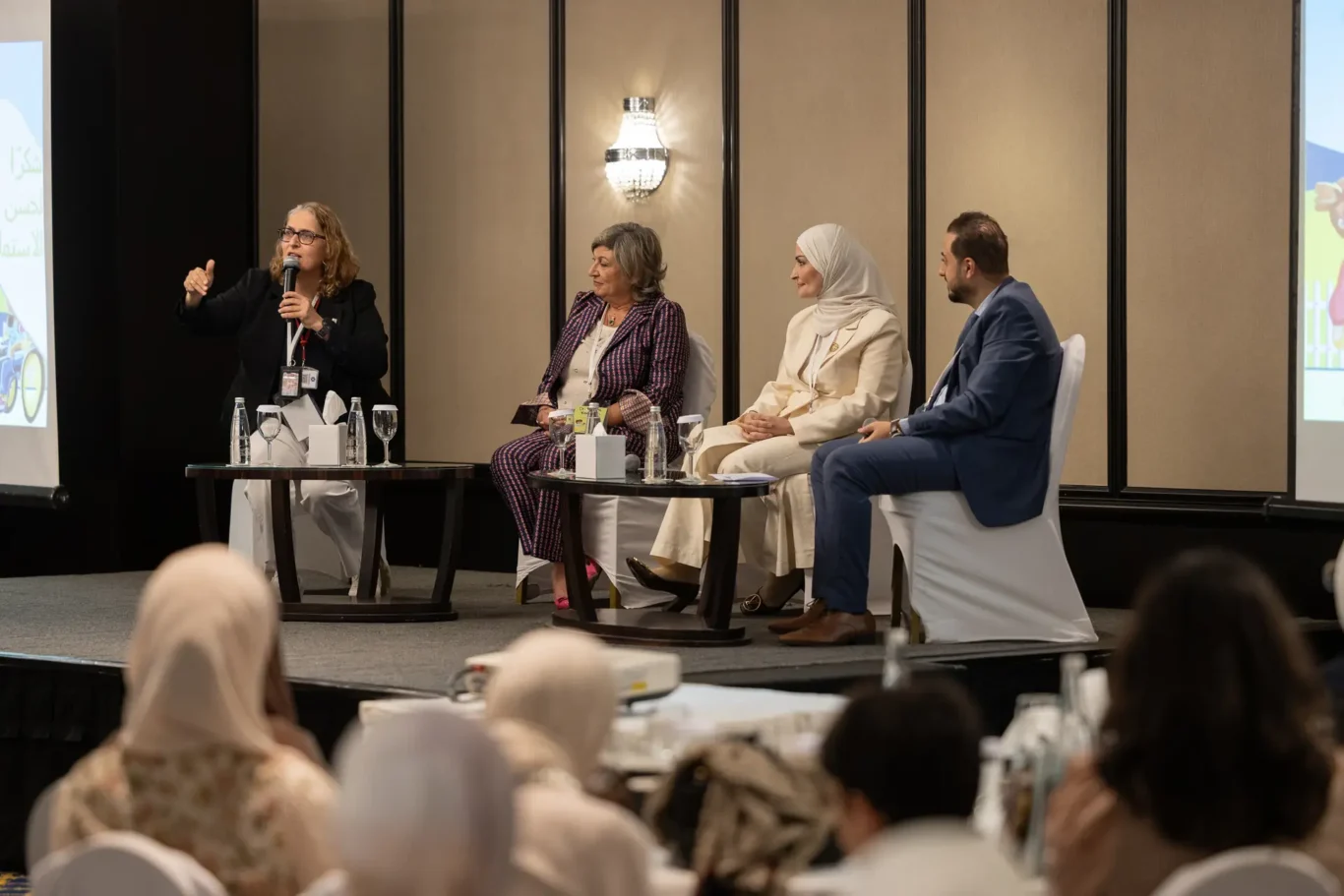 Panellist speaking into a microphone on stage with three other panellists seated beside her. Corporate event photography by MRPS for My Arabic Publishing at Movenpick Grand Al Bustan, Dubai.