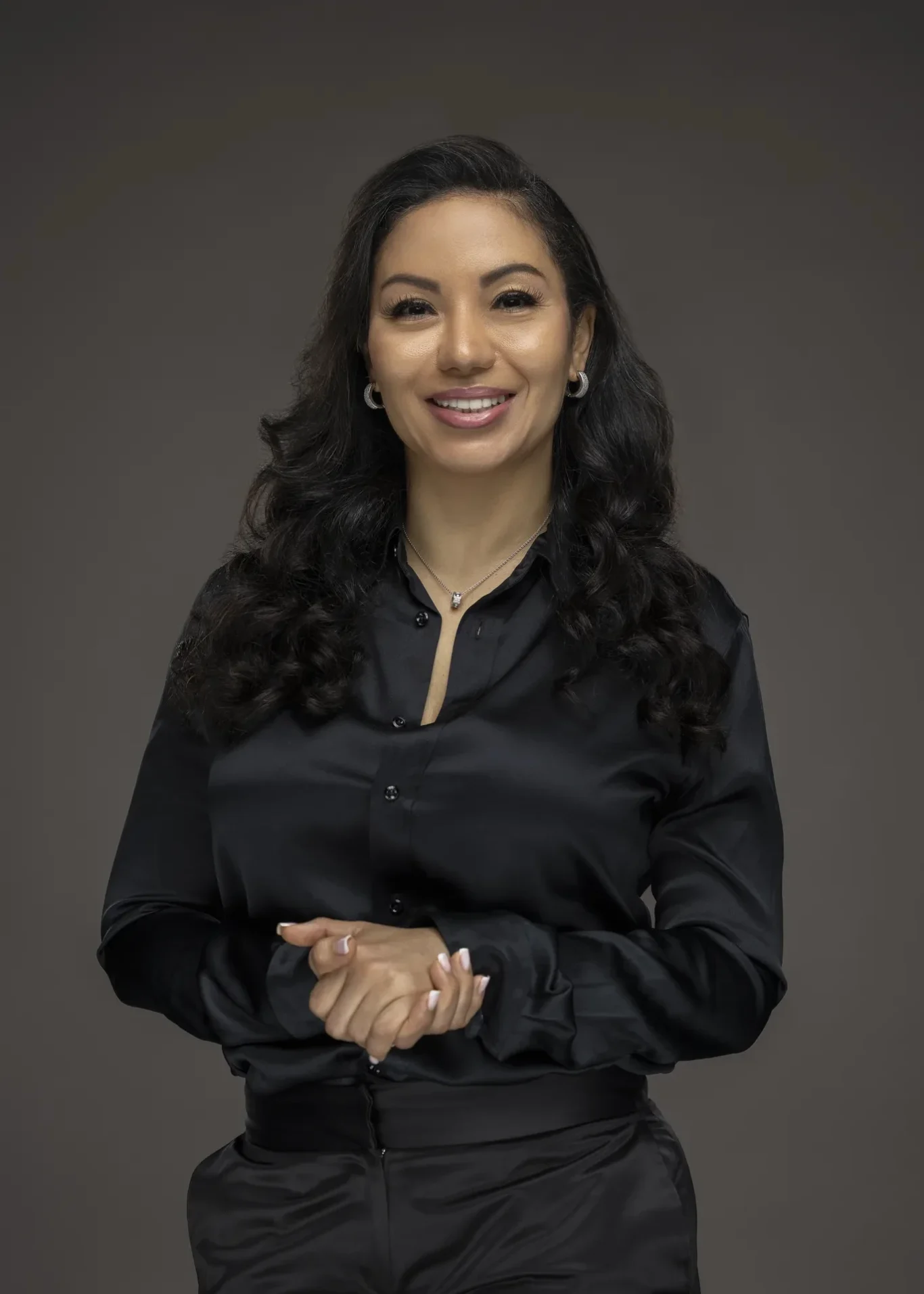 Sandra in a black satin shirt with hands together on a grey backdrop. Business profile and LinkedIn portrait photography by MRPS in Dubai.
