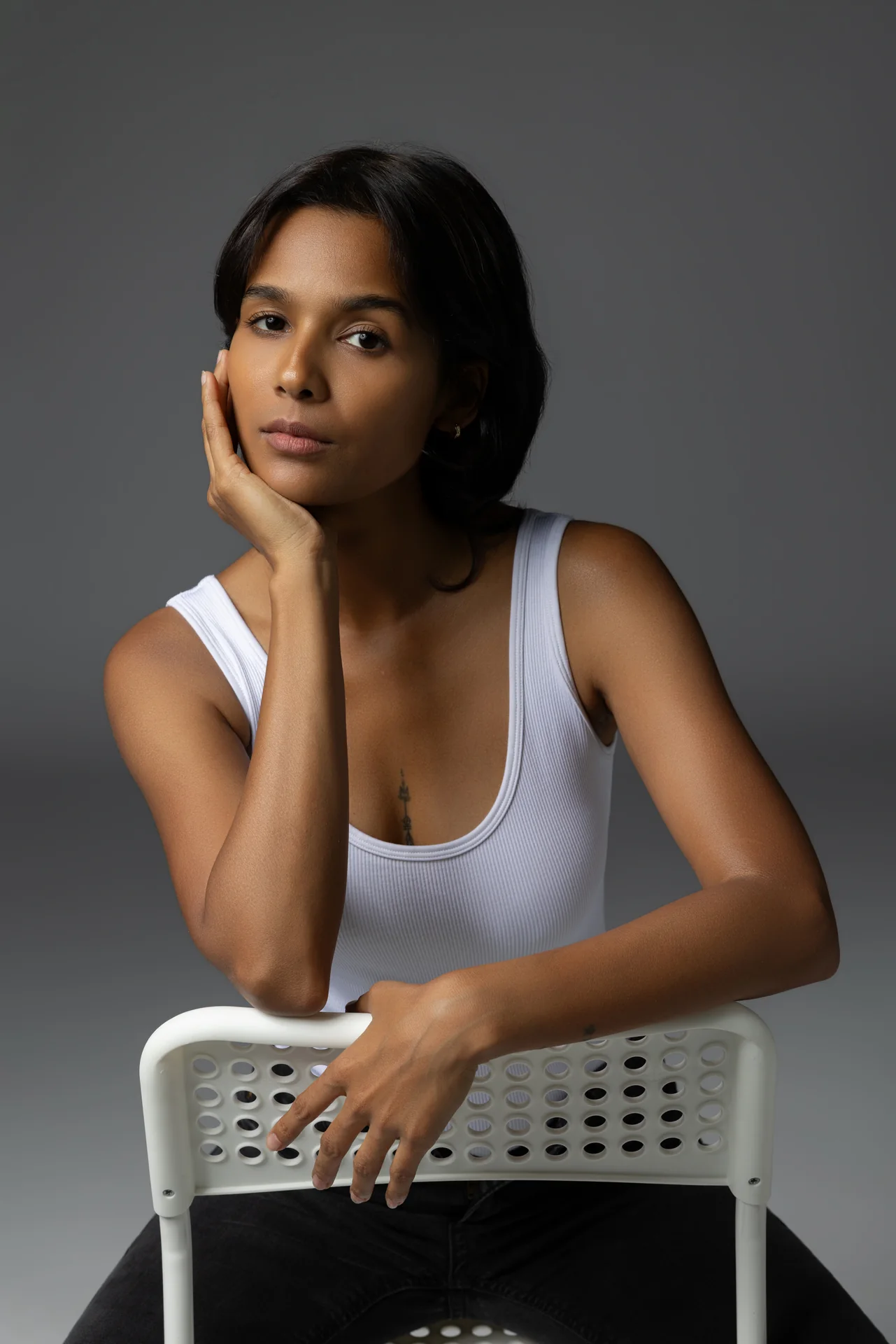 Mia leaning on the back of a white chair with her hand on her cheek, wearing a white tank top, in a studio portfolio portrait.