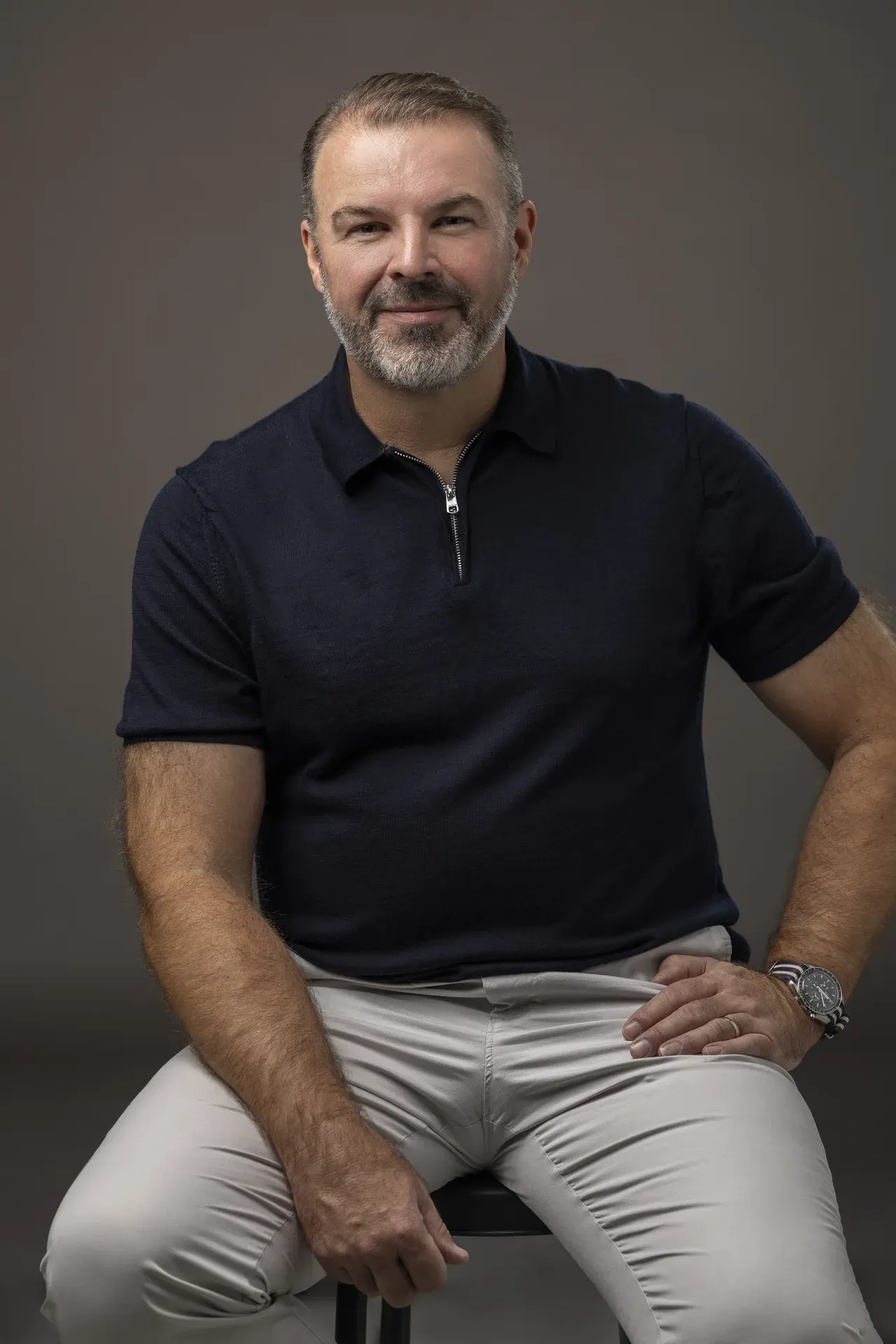 Rik Williams seated on a stool in a navy polo and light chinos on a grey backdrop. Corporate portrait photography by MRPS in Dubai, photographed by Nastaran.