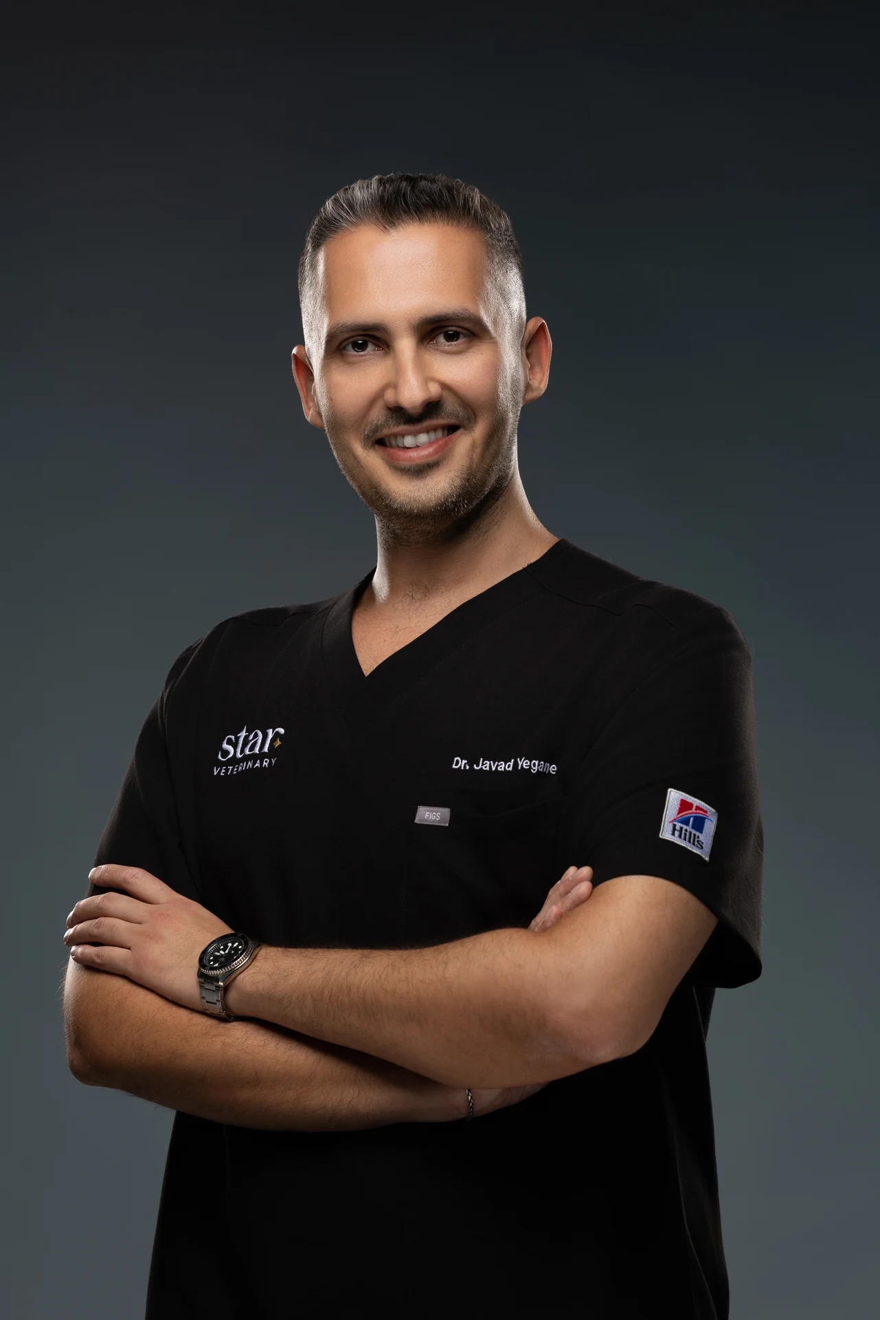 Veterinarian headshot in black clinic scrubs with arms crossed and a warm smile. Photographed at the MRPS studio in Business Bay, Dubai.