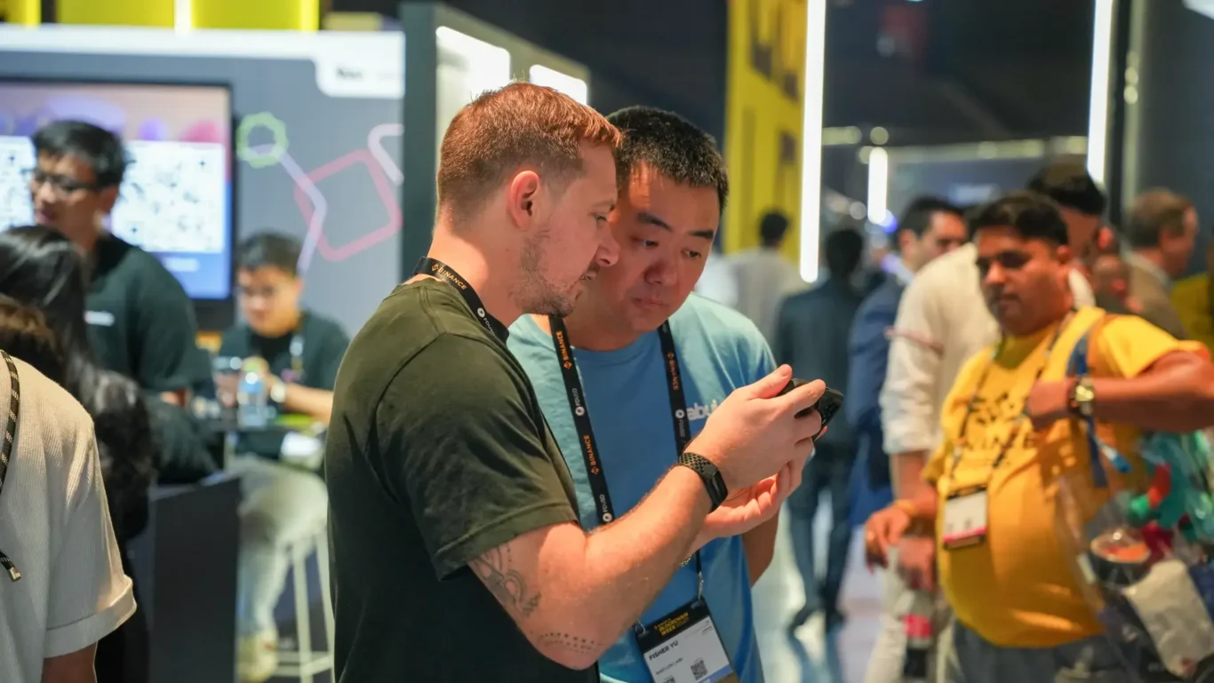 Visitors comparing phones and networking at the booth. Photographed by MRPS for Eventflare at Binance Blockchain Week 2025, Coca-Cola Arena, Dubai.