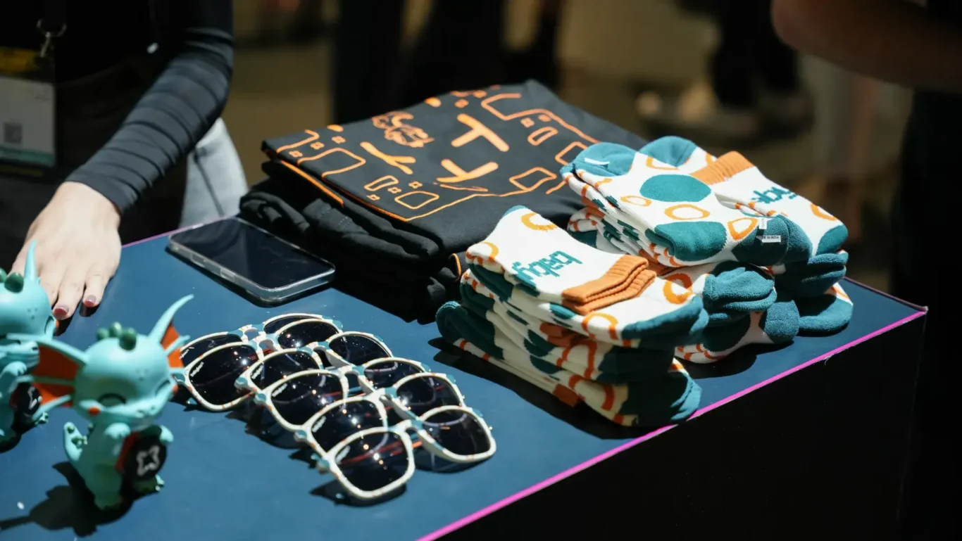 Babylon branded socks, sunglasses and collectibles on the booth counter. Photographed by MRPS for Eventflare at Binance Blockchain Week 2025, Coca-Cola Arena, Dubai.