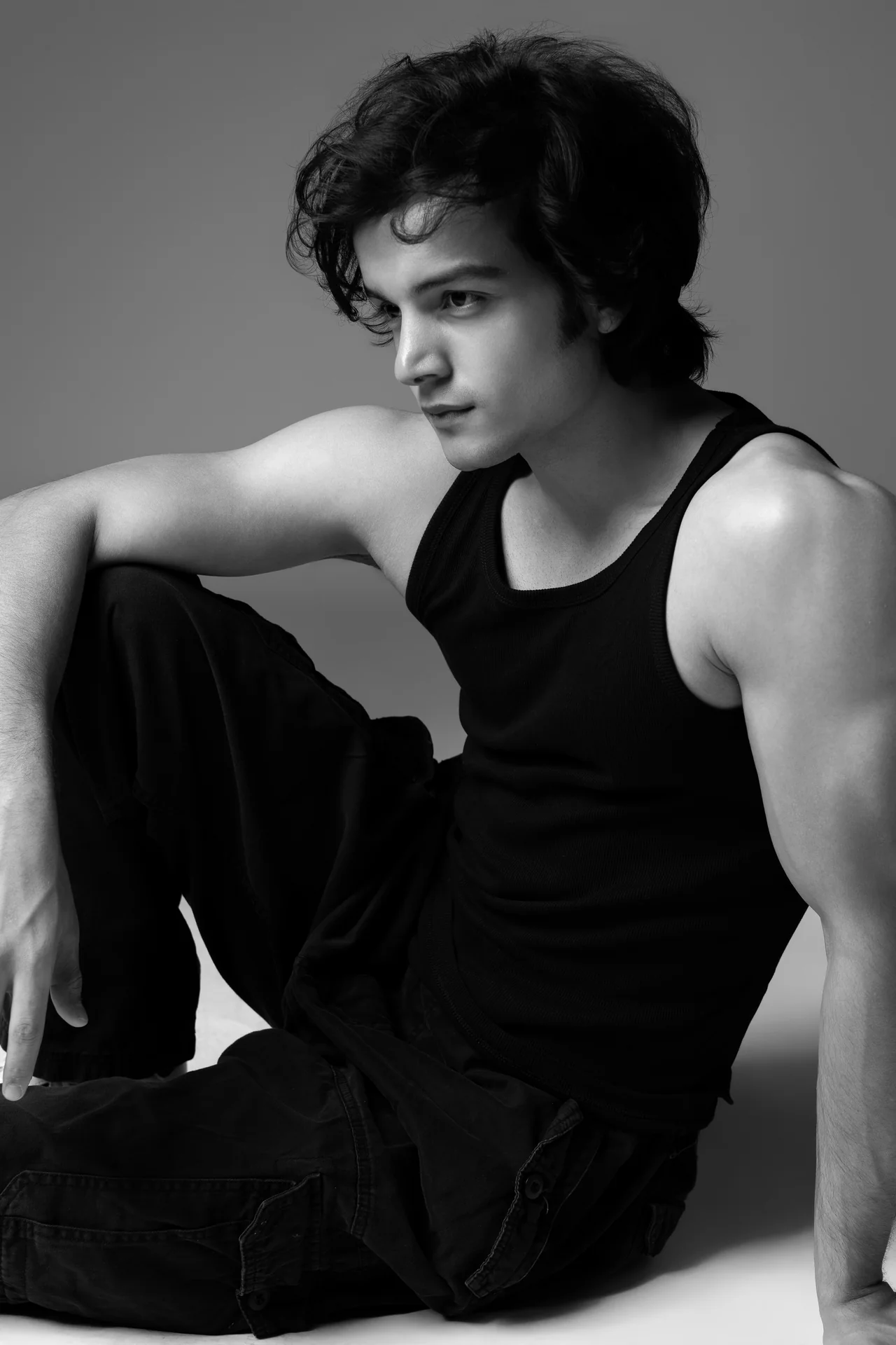 Black and white close portrait of Arsh in a black tank top, seated on the studio floor.