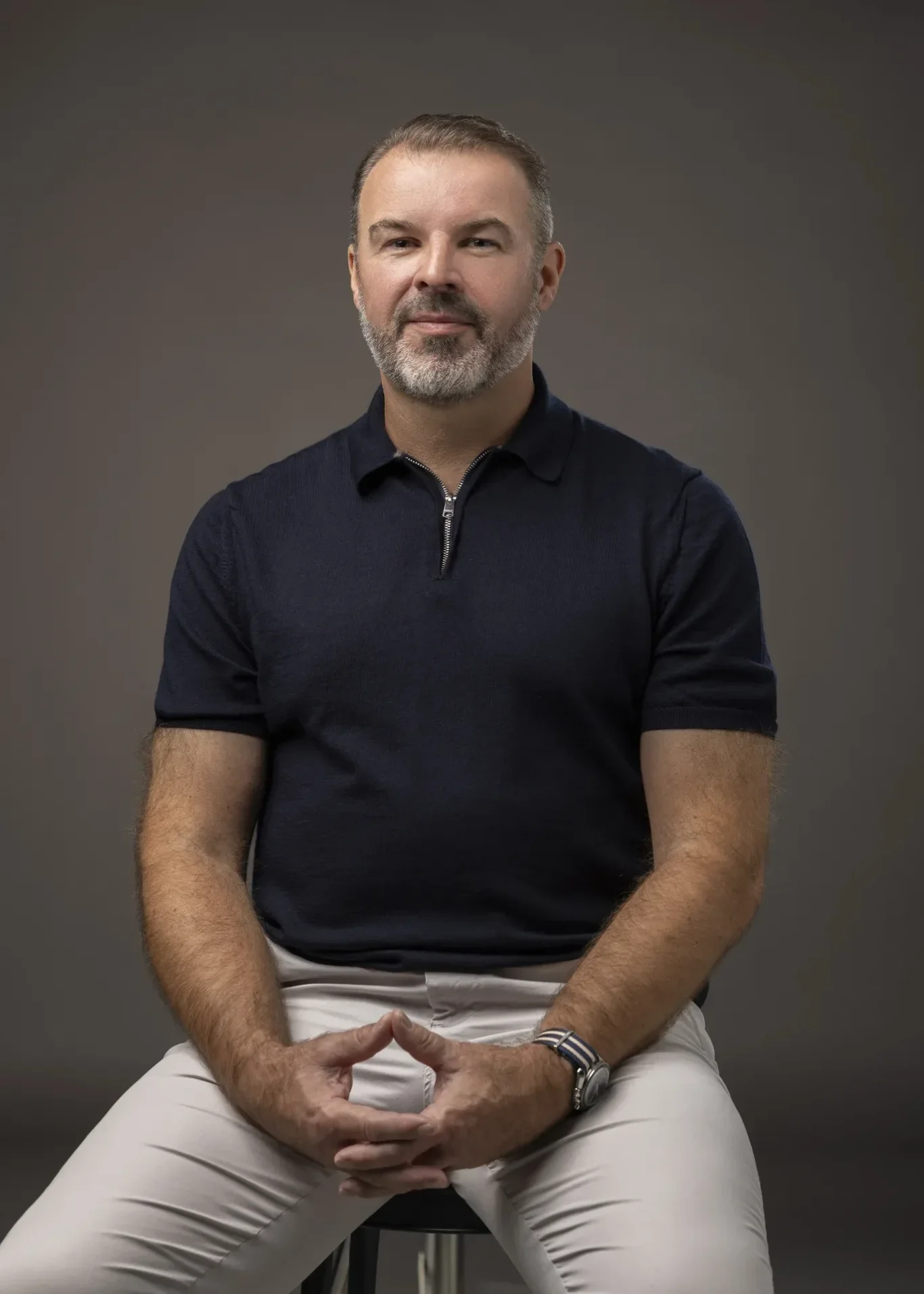 Seated portrait of Rik Williams in a navy polo with hands together, grey backdrop. Corporate portrait photography by MRPS in Dubai, photographed by Nastaran.