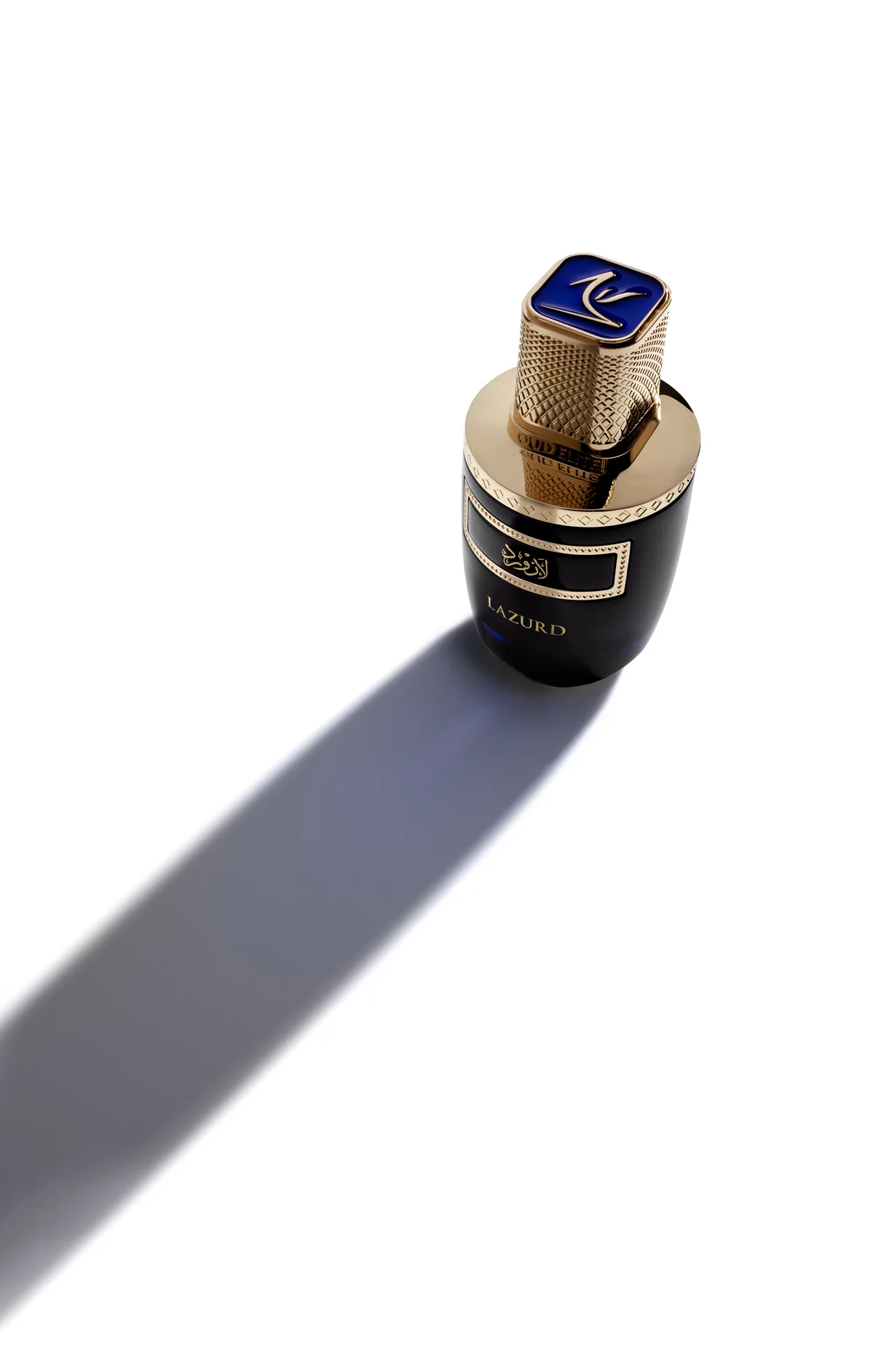 High-angle shot of the Oud Elite Lazurd perfume bottle showing its gold cap with blue emblem and a long soft shadow.