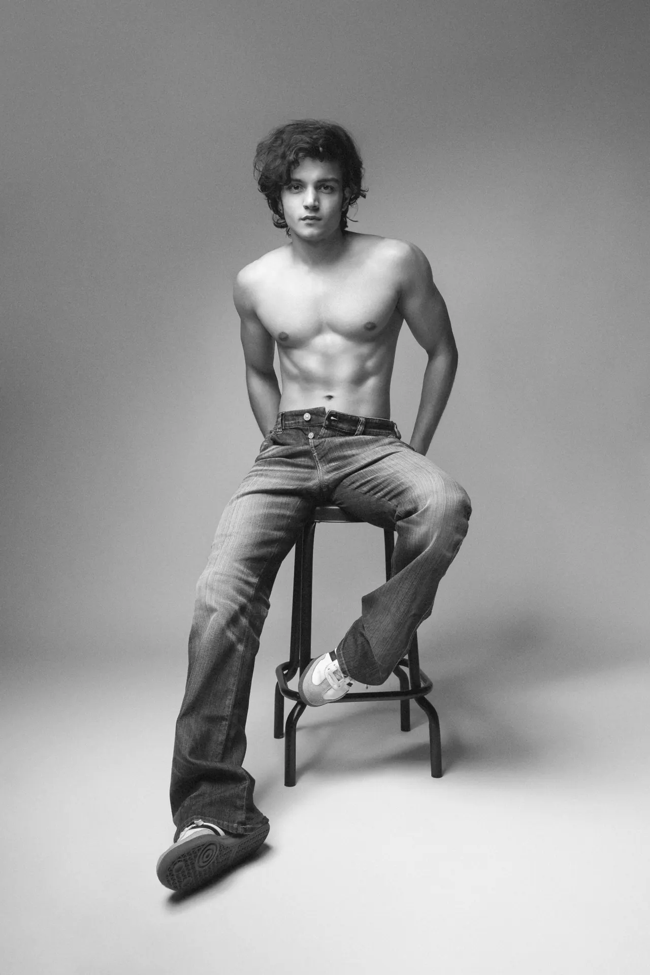 Black and white shot of Arsh shirtless in faded jeans, seated on a studio stool.