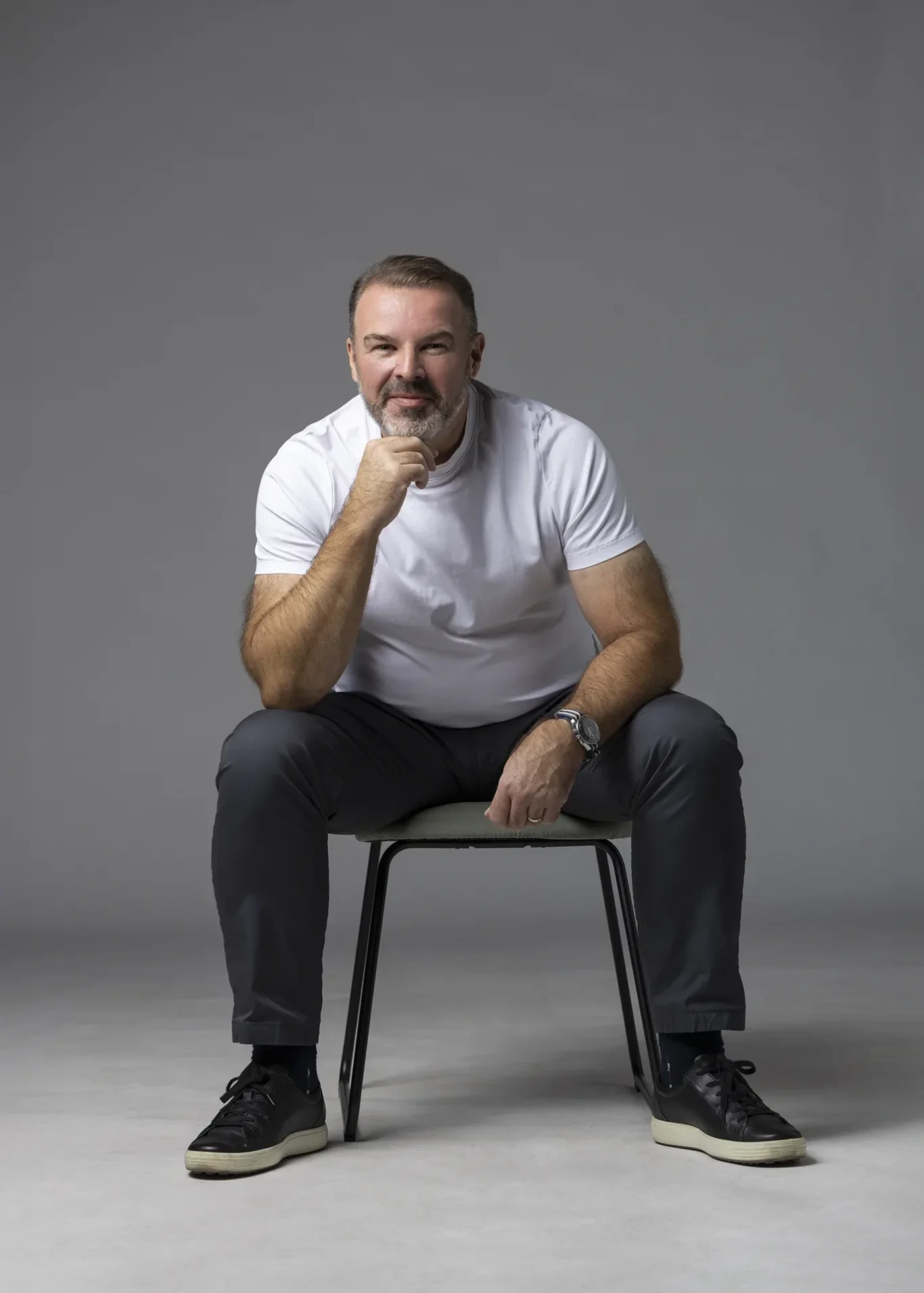 Rik Williams seated in a white t-shirt with a hand on his chin, light grey backdrop. Corporate portrait photography by MRPS in Dubai, photographed by Nastaran.