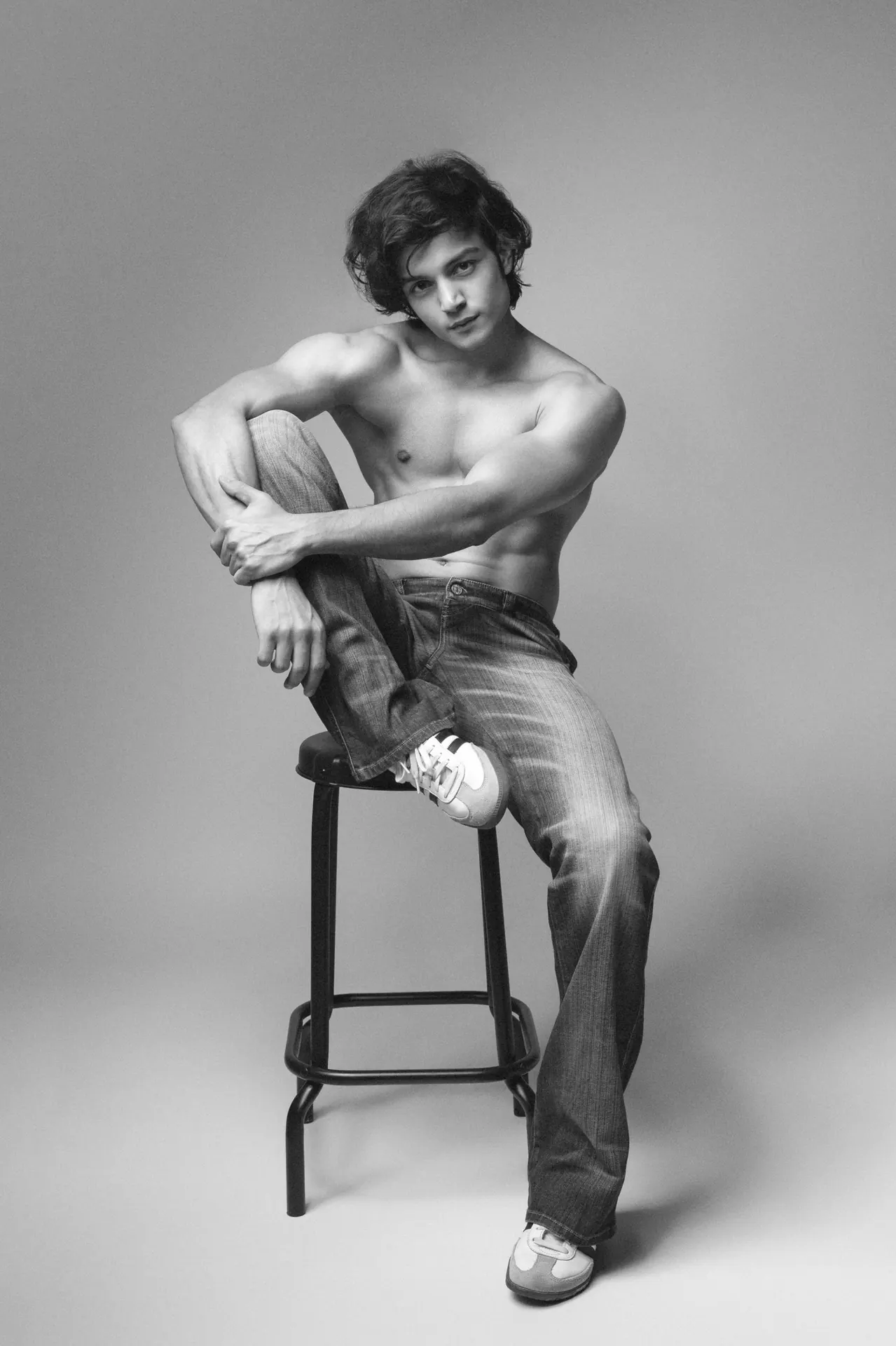 Black and white shot of Arsh shirtless on a studio stool with one knee raised, looking at the camera.