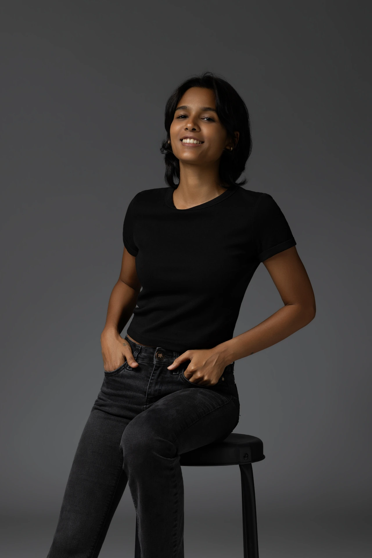 Smiling portrait of Mia seated on a stool in a black t-shirt and black jeans.