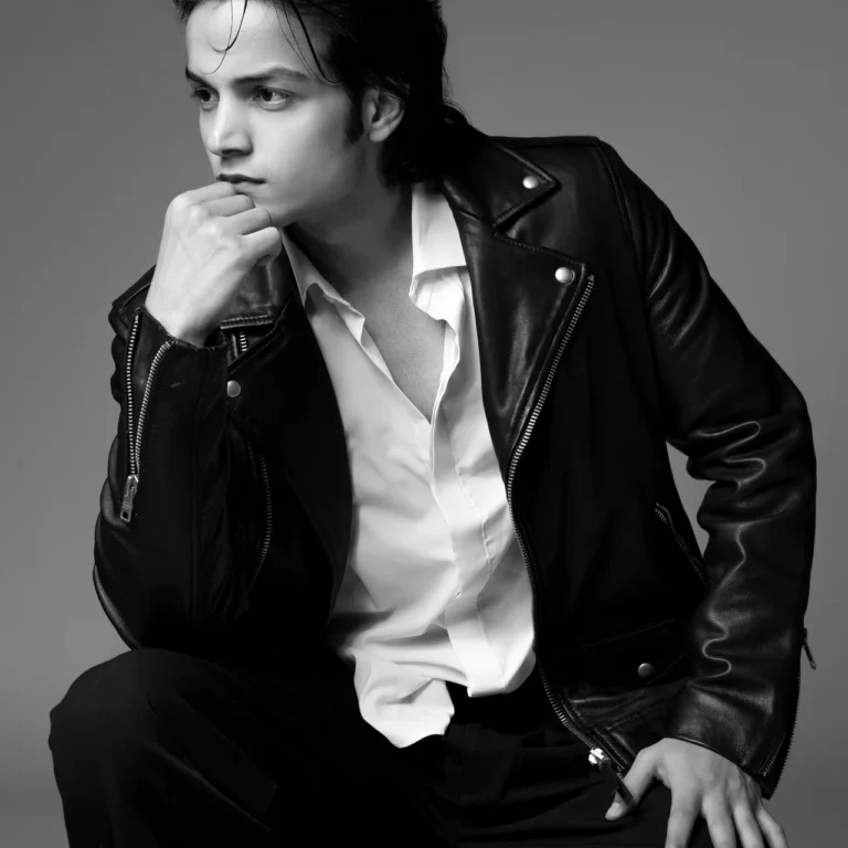 Black and white editorial portrait of Arsh in a leather jacket and white shirt, seated with his hand on his chin.