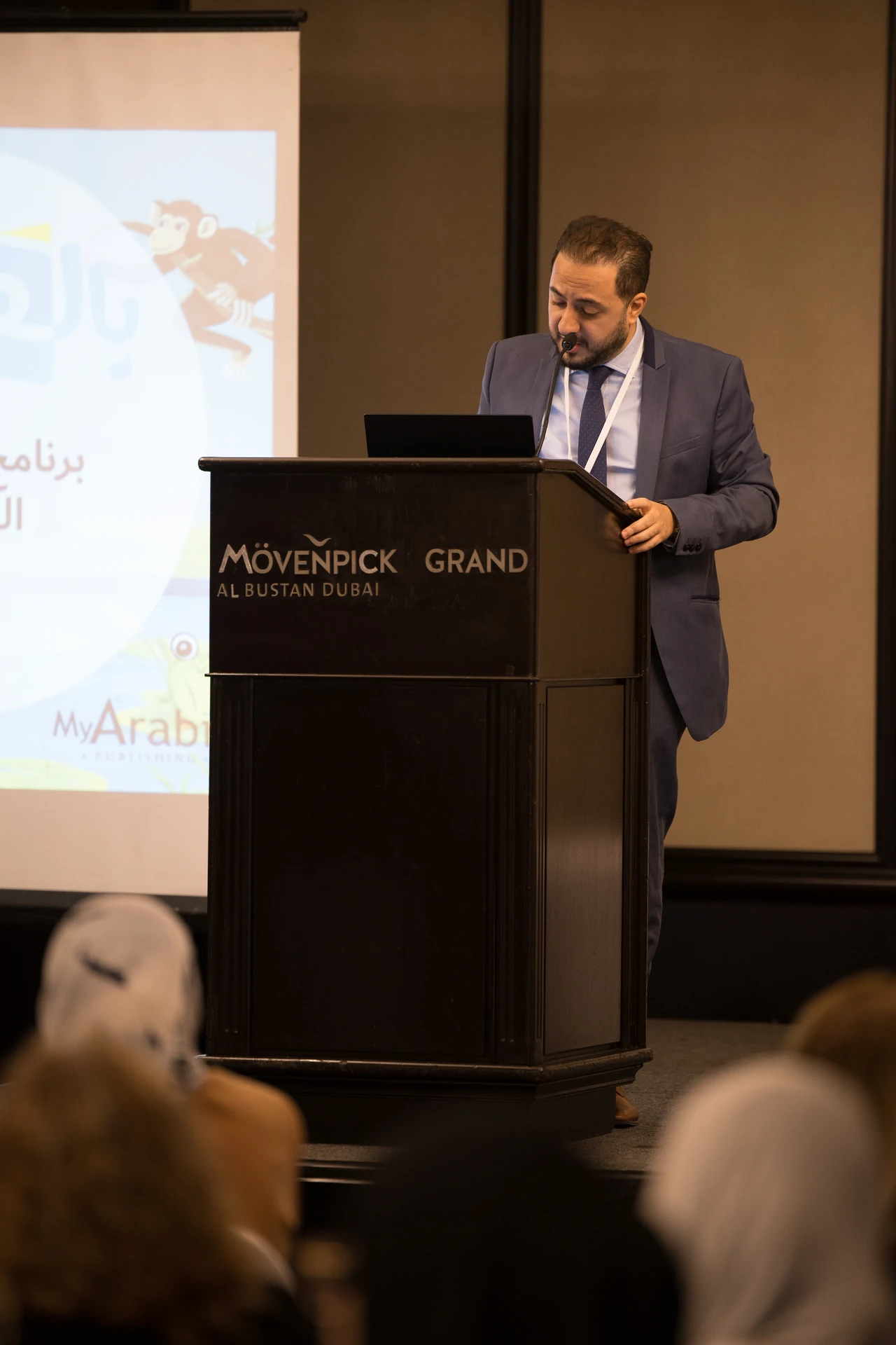 Presenter at the podium with the My Arabic slides on the screen behind him. Corporate event photography by MRPS for My Arabic Publishing at Movenpick Grand Al Bustan, Dubai.