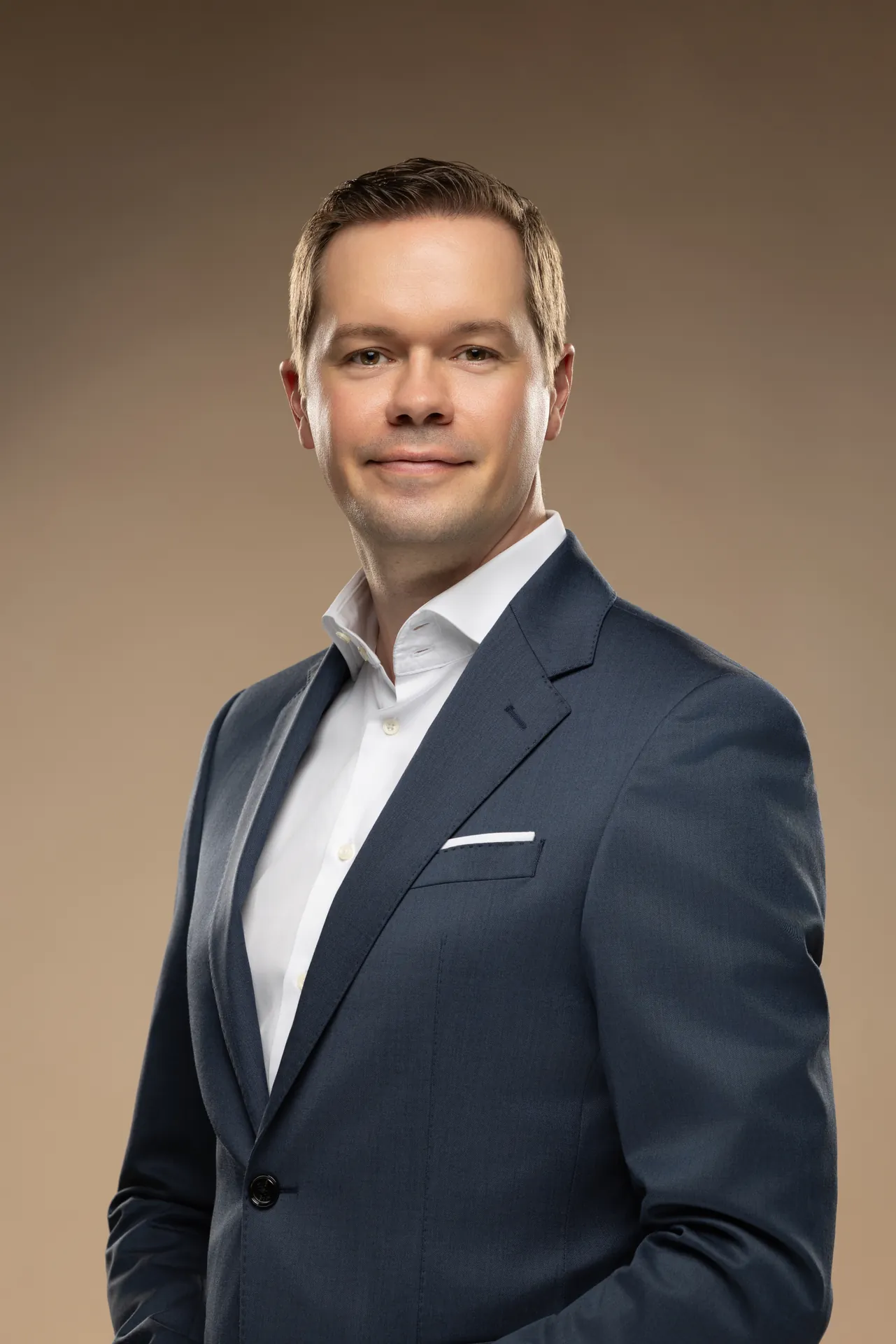 Website profile portrait of an executive in a navy suit with an open collar on a beige studio backdrop.