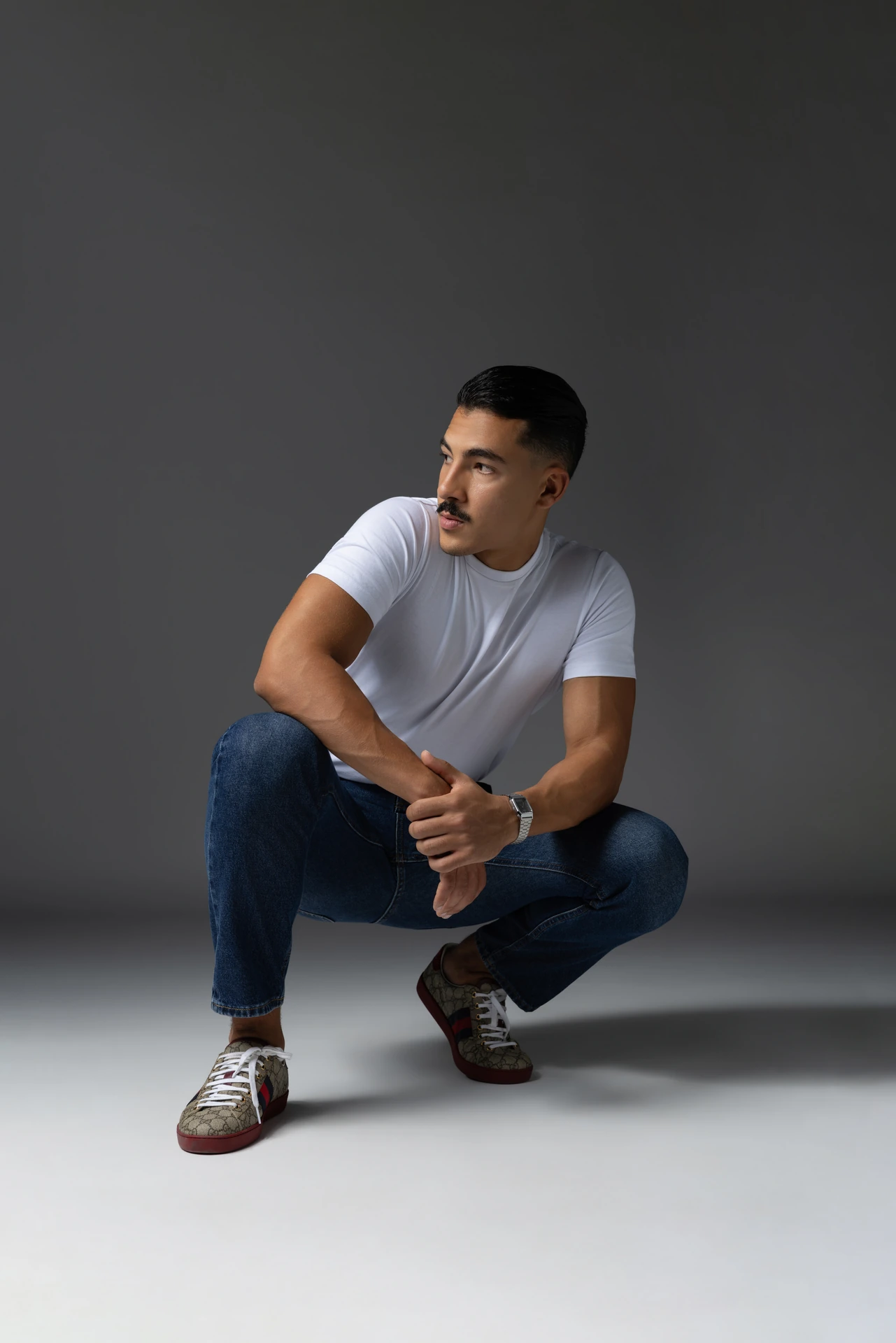 Crouching pose in a white t-shirt and jeans on a grey backdrop. Male modeling portfolio photoshoot by MRPS in Dubai, photographed by Nastaran.