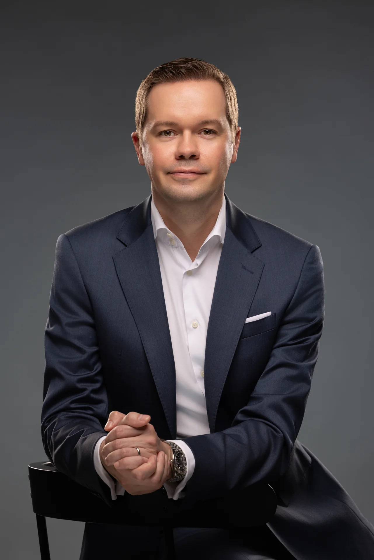 Seated corporate portrait of an executive in a navy suit leaning forward with hands clasped, grey backdrop.