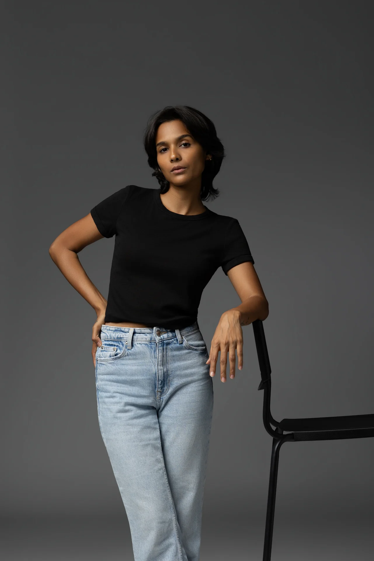 Mia standing beside a black chair in a black t-shirt and light blue jeans, hand on hip.