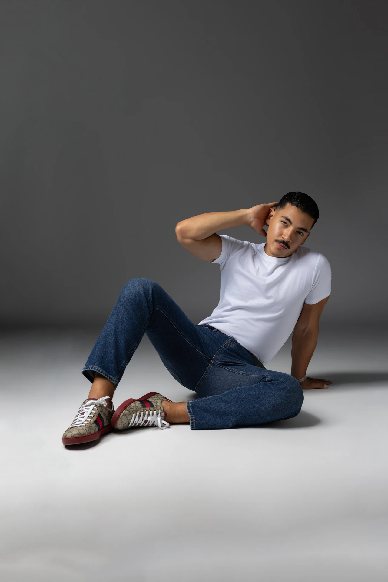 Sitting on the studio floor in a white t-shirt and jeans, hand behind the head. Male modeling portfolio photoshoot by MRPS in Dubai, photographed by Nastaran.